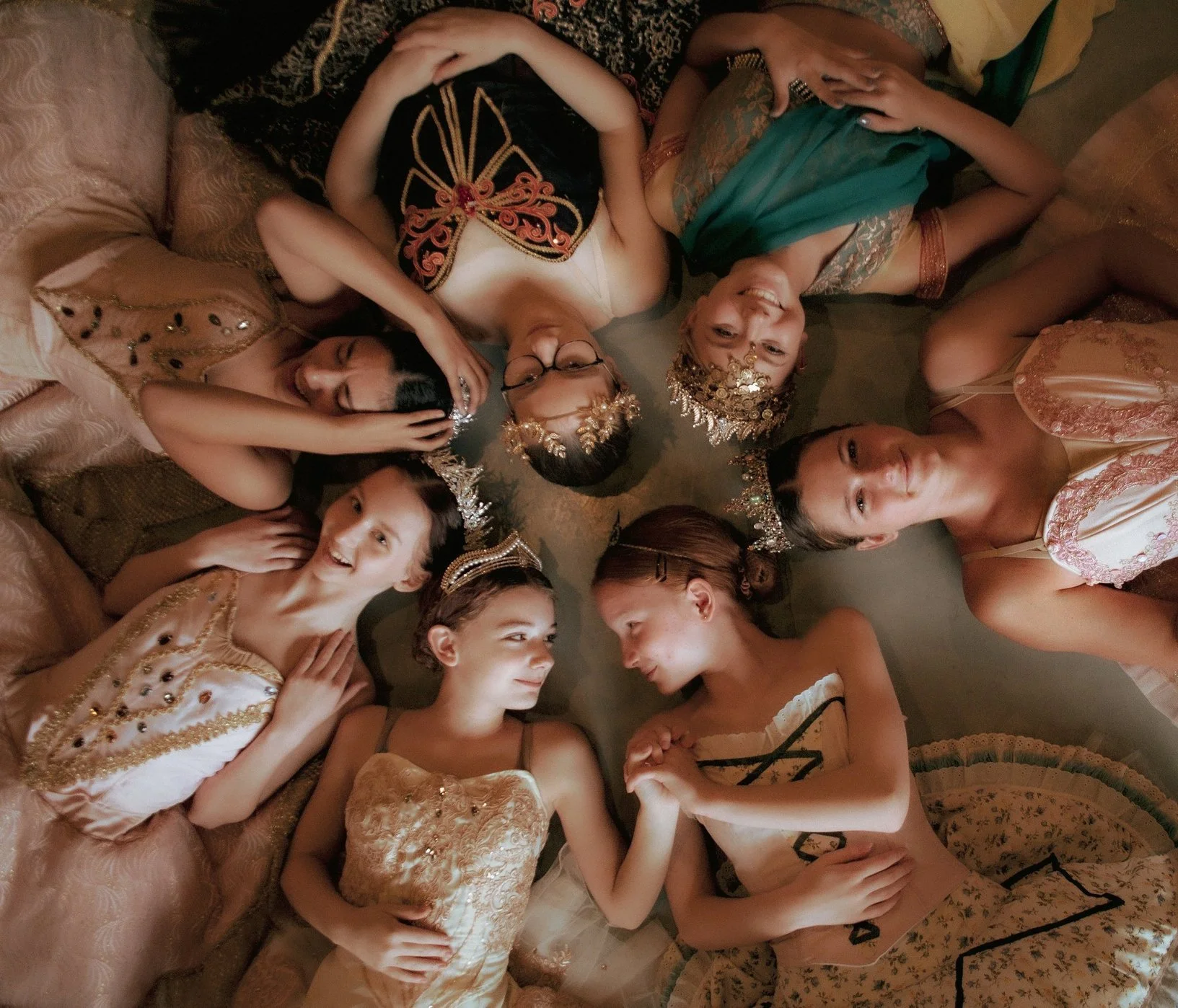 Group of women lying on the floor in a circle, wearing fancy dresses and tiaras, smiling and interacting with each other.