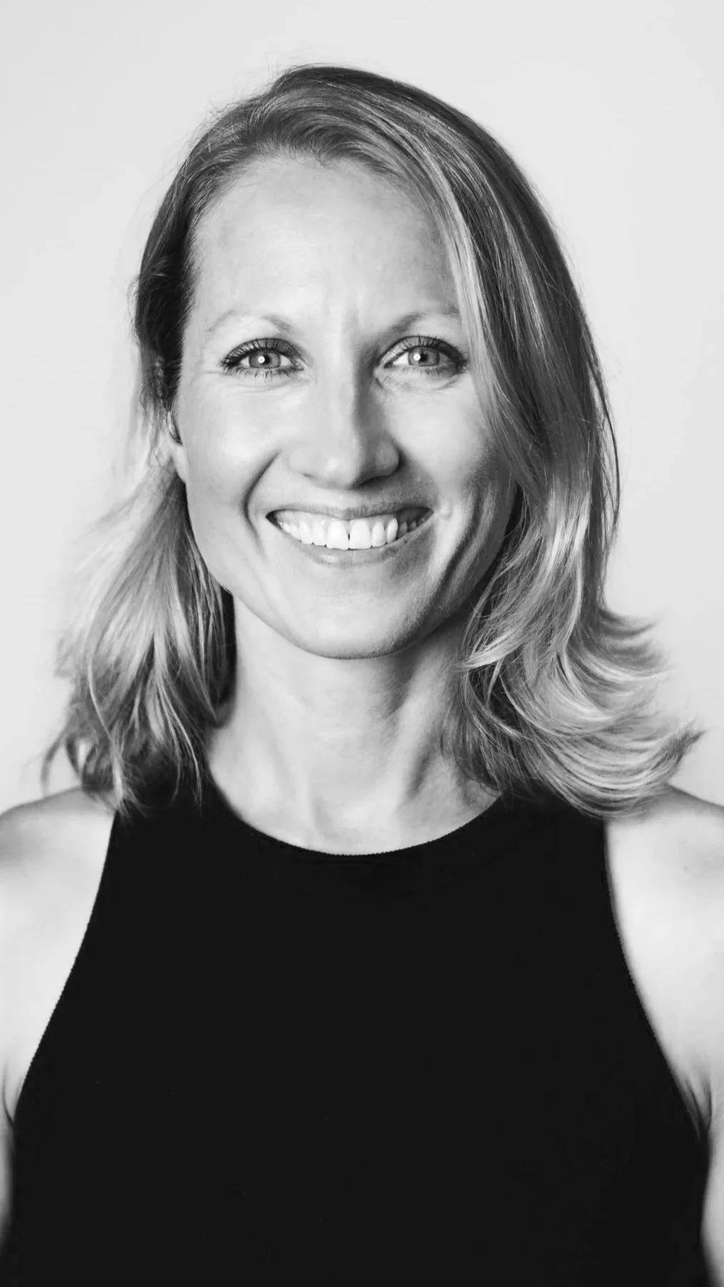 Black and white portrait of a woman smiling, with shoulder-length hair, wearing a sleeveless black top.