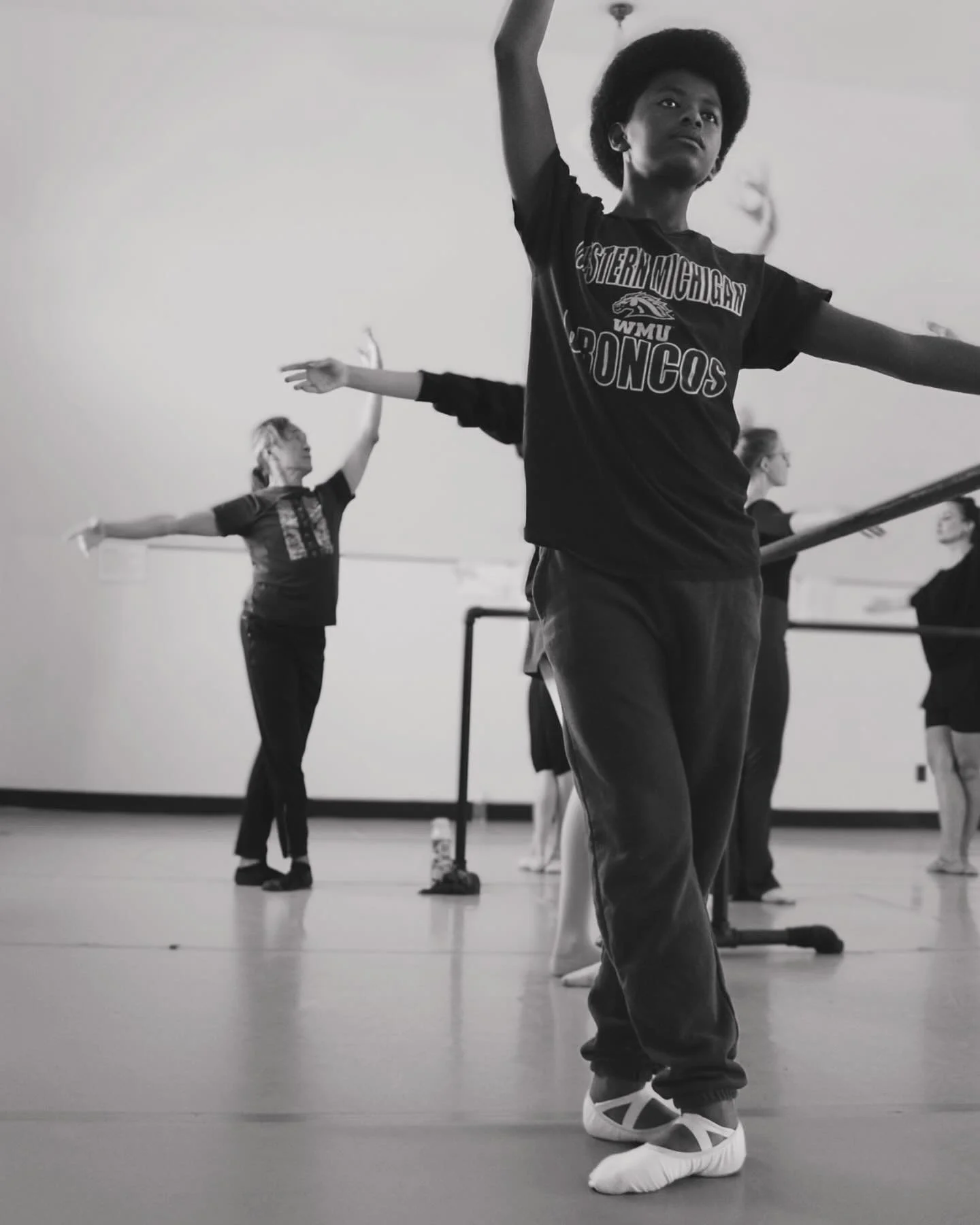 A group of people in a dance class practicing ballet or modern dance in a studio. The focus is on a young woman in the foreground with arms outstretched, wearing a dark T-shirt with sports team graphics and loose pants, balanced with dance shoes. Other participants are in the background, also stretching or practicing dance moves.