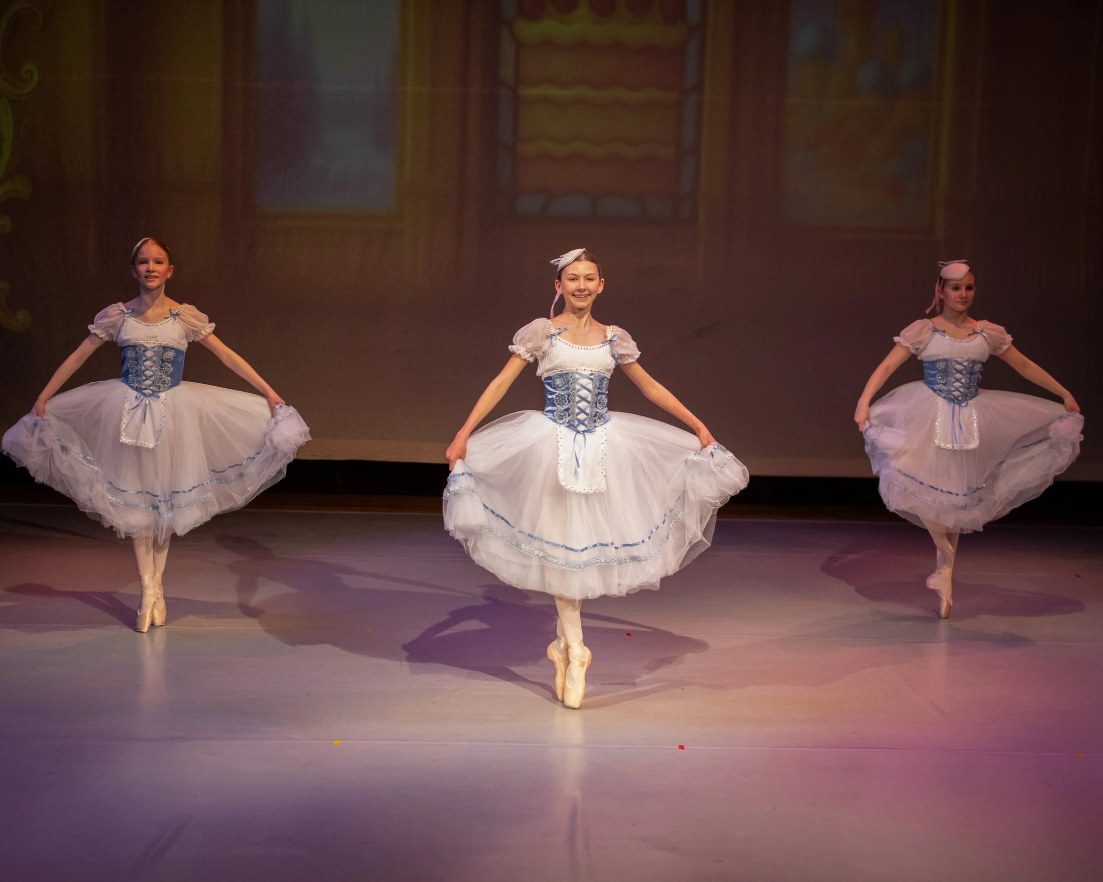 Three ballerinas performing on stage, wearing white tutus with blue accents and matching headpieces, dancing en pointe against a dimly lit background.
