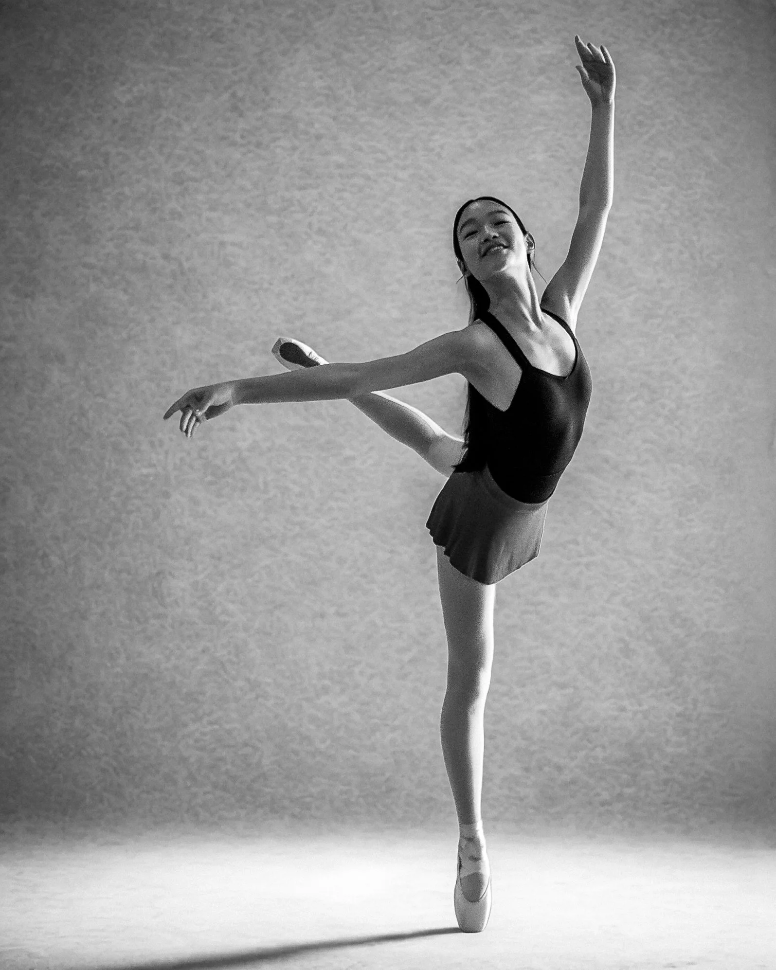 A ballerina in a black leotard and skirt balancing on one foot with her arms raised in a dance pose, black and white photo