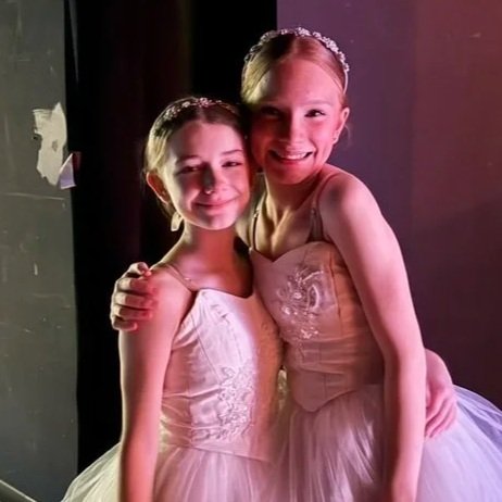 Two young girls in ballet tutus smiling together, one with her arm around the other, standing in a dimly lit room.