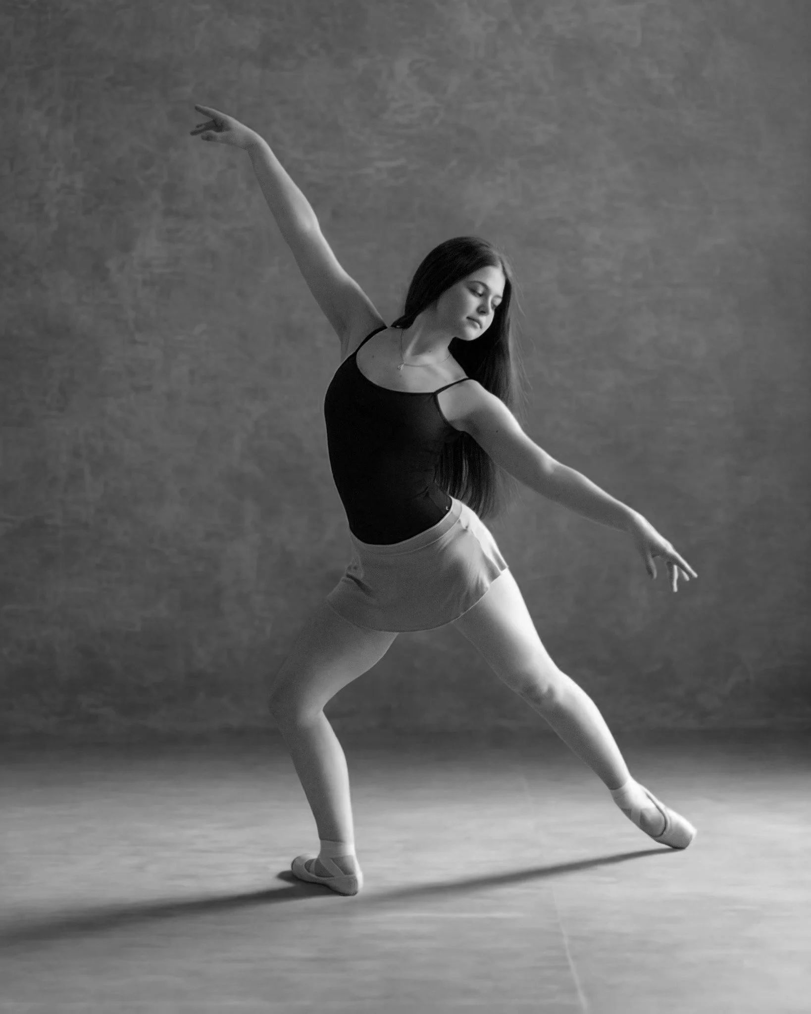 A ballerina practicing ballet, standing on one leg with the other leg extended behind her, arms outstretched, wearing a leotard and ballet slippers on a dance floor.