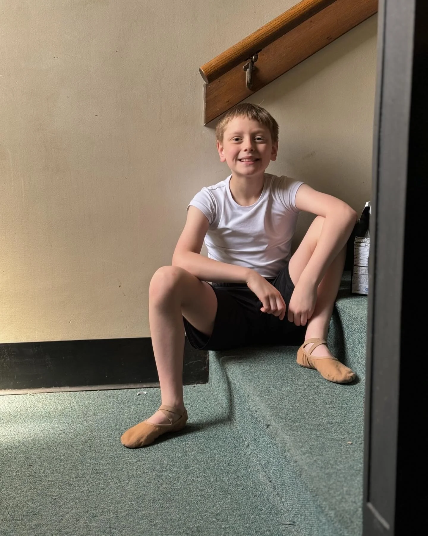 A young boy sitting on a green carpeted staircase, smiling, wearing a white t-shirt, black shorts, and tan ballet flats, with a beige wall and wooden handrail in the background.