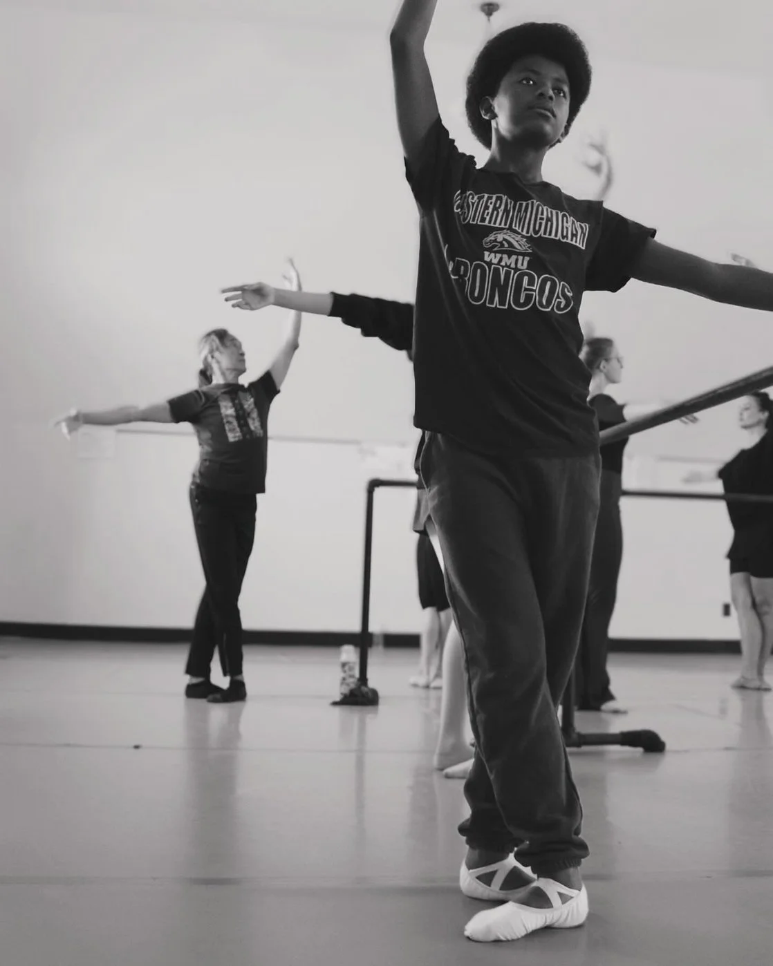 Young ballet dancers practicing at a dance studio, with one boy in the foreground wearing a Western Michigan Broncos shirt, balancing on his toes with arms raised, and women in the background stretching and practicing at the ballet barre.