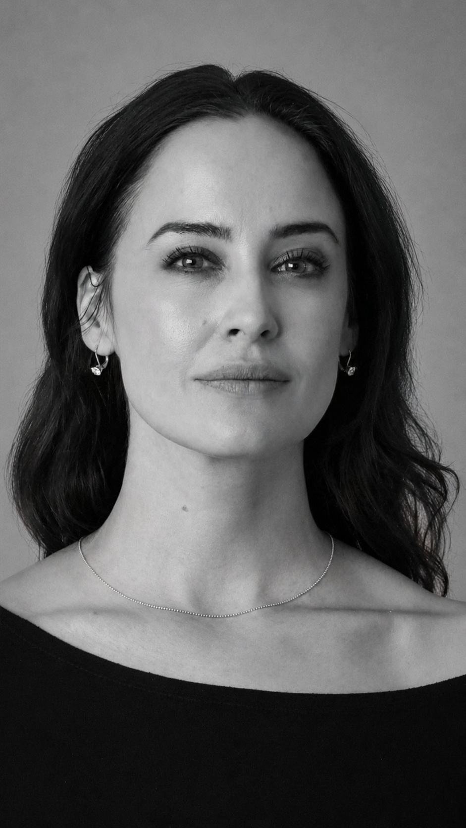 Black and white portrait of a woman with dark hair, wearing earrings and a thin necklace, looking directly at the camera with a subtle smile.