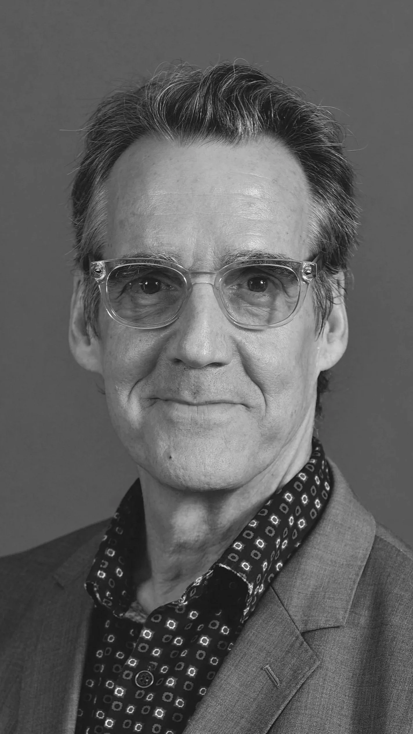 Black and white portrait of an older man with glasses, styled hair, wearing a patterned shirt and a blazer.