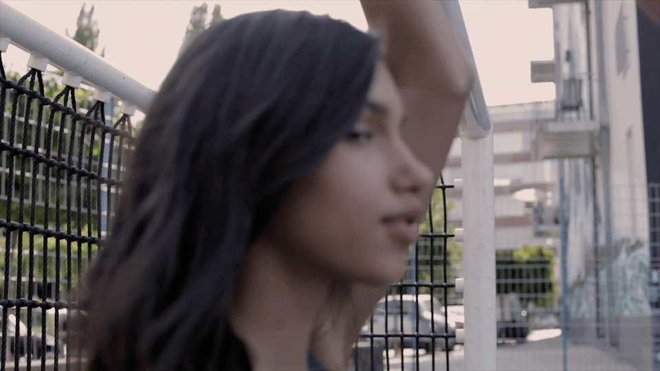 A woman with long dark hair standing near a fence on an outdoor sports field or track.