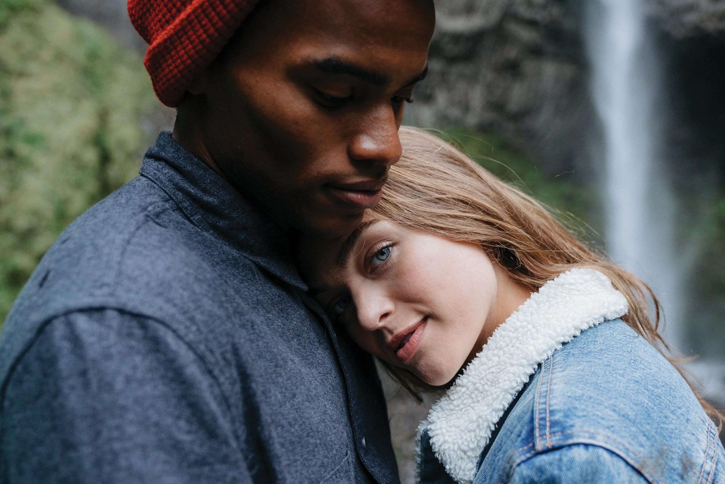 A young couple embracing each other outdoors, with a blurred waterfall and greenery in the background.