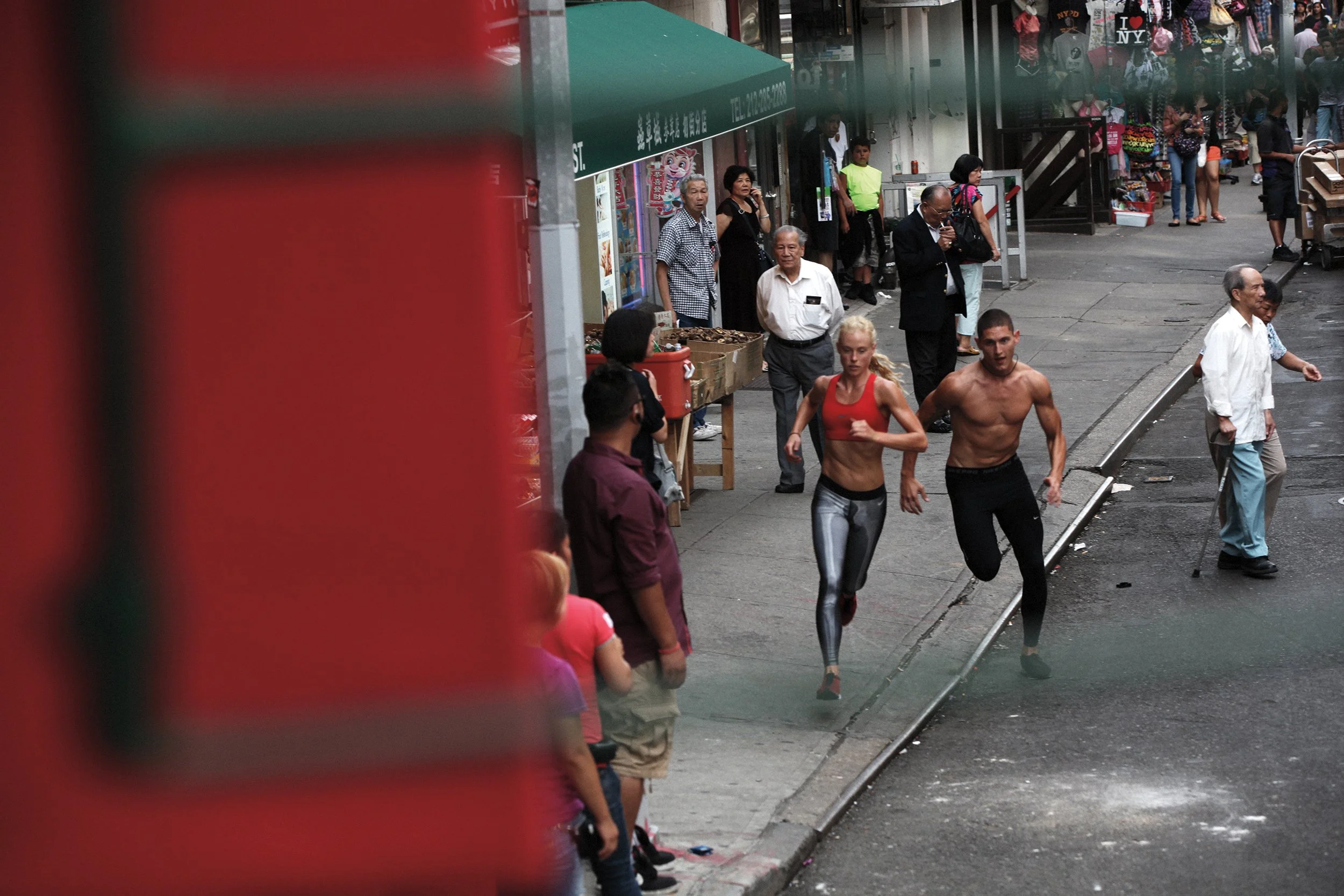 Two people jogging on a city sidewalk, with onlookers and storefronts in the background.