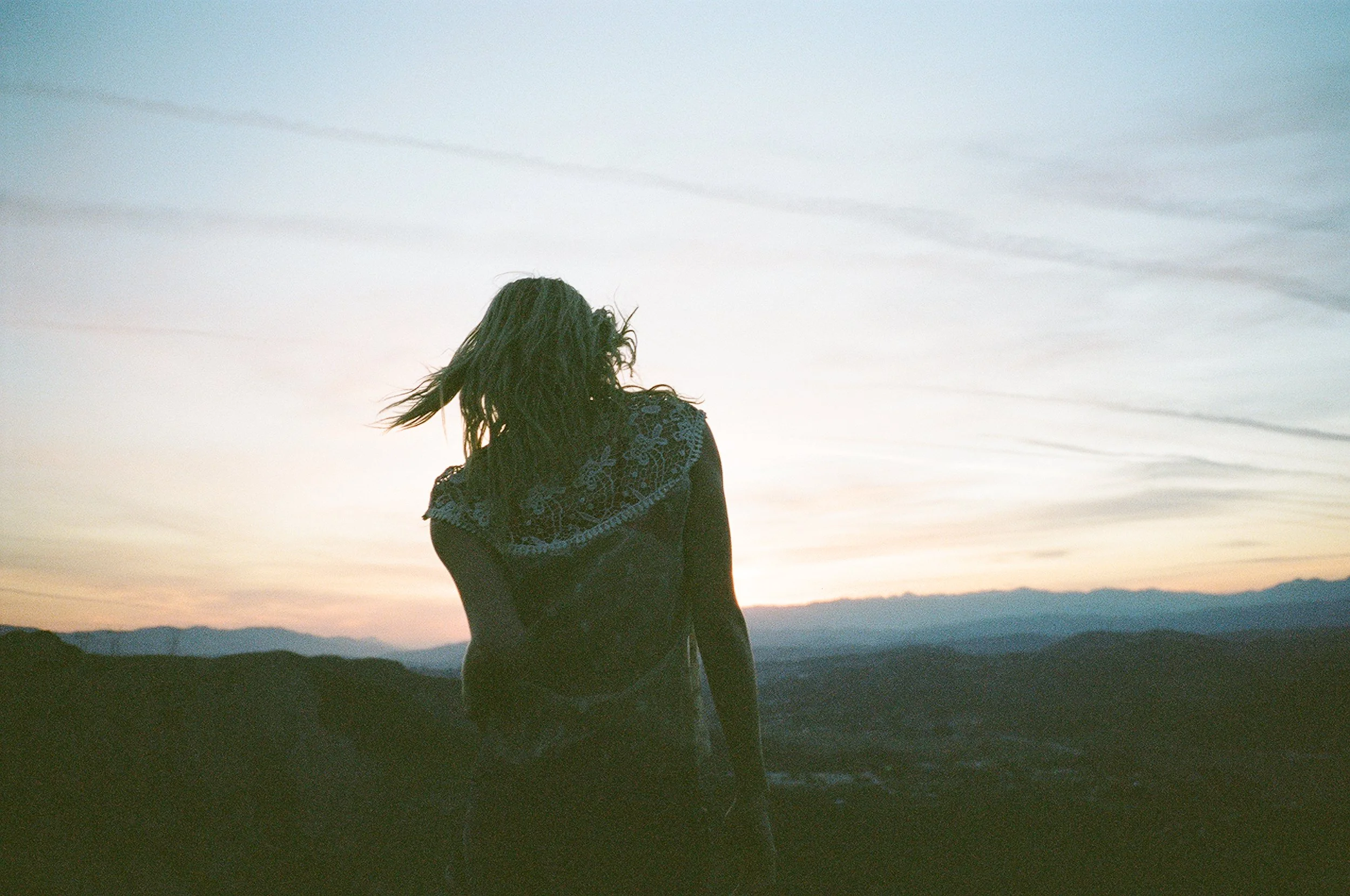 A woman with dark hair standing outdoors on a hill at sunset, looking away from the camera with mountains and a colorful sky in the background.
