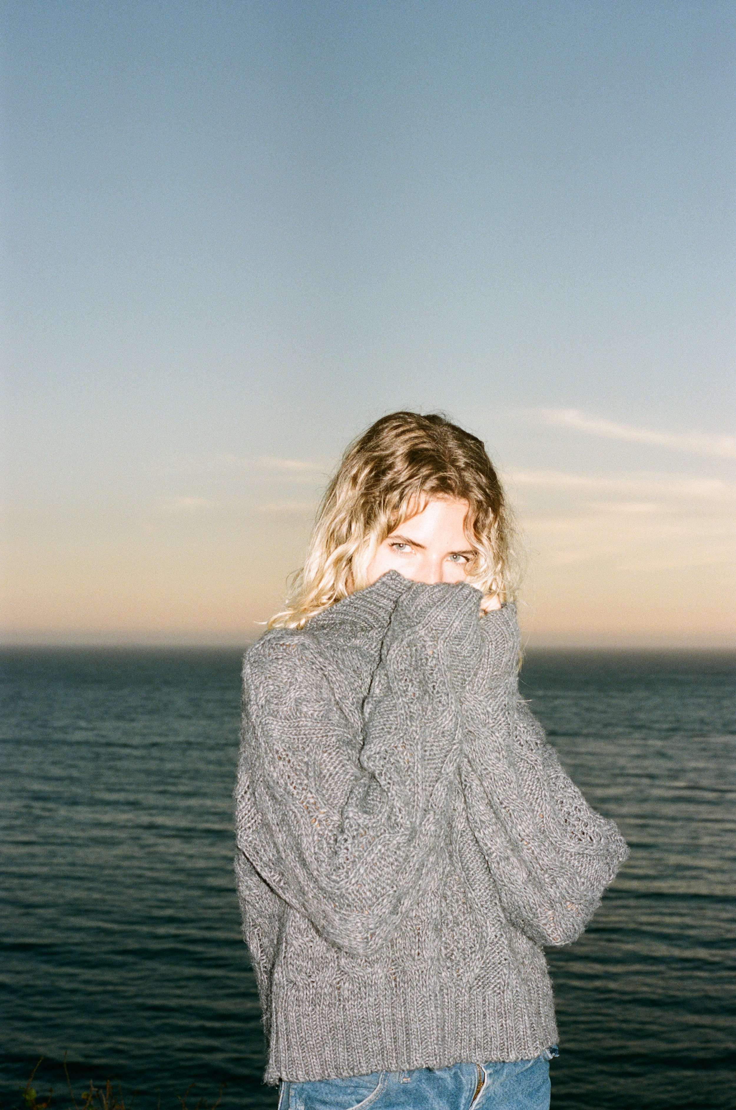 A woman with blonde, curly hair wearing a gray sweater covers her mouth with the sweater while standing outdoors near a large body of water, with a clear blue sky in the background.