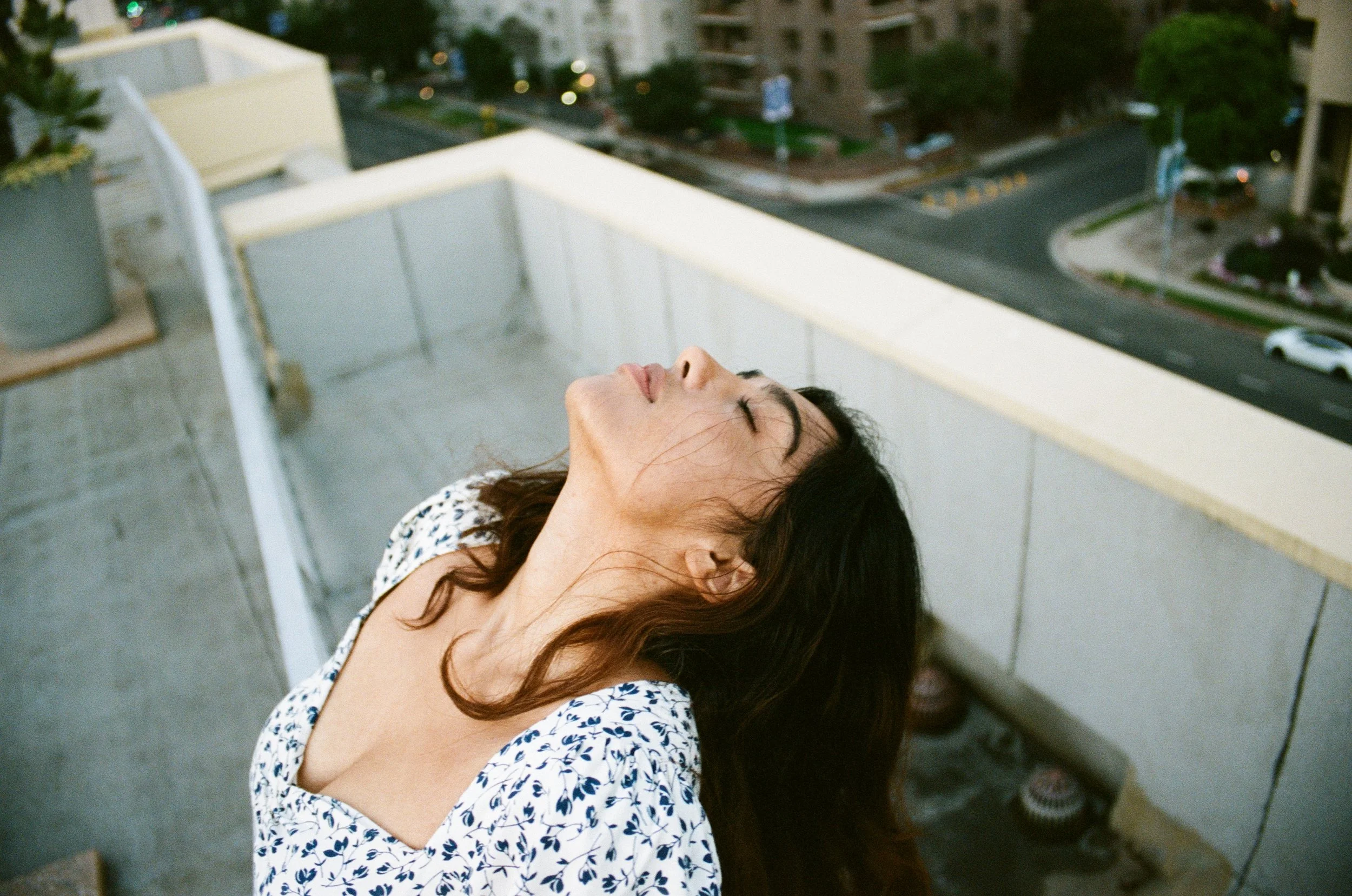 A woman with dark hair and a patterned top tilts her head back with eyes closed on a balcony, overlooking a city street with cars and buildings.