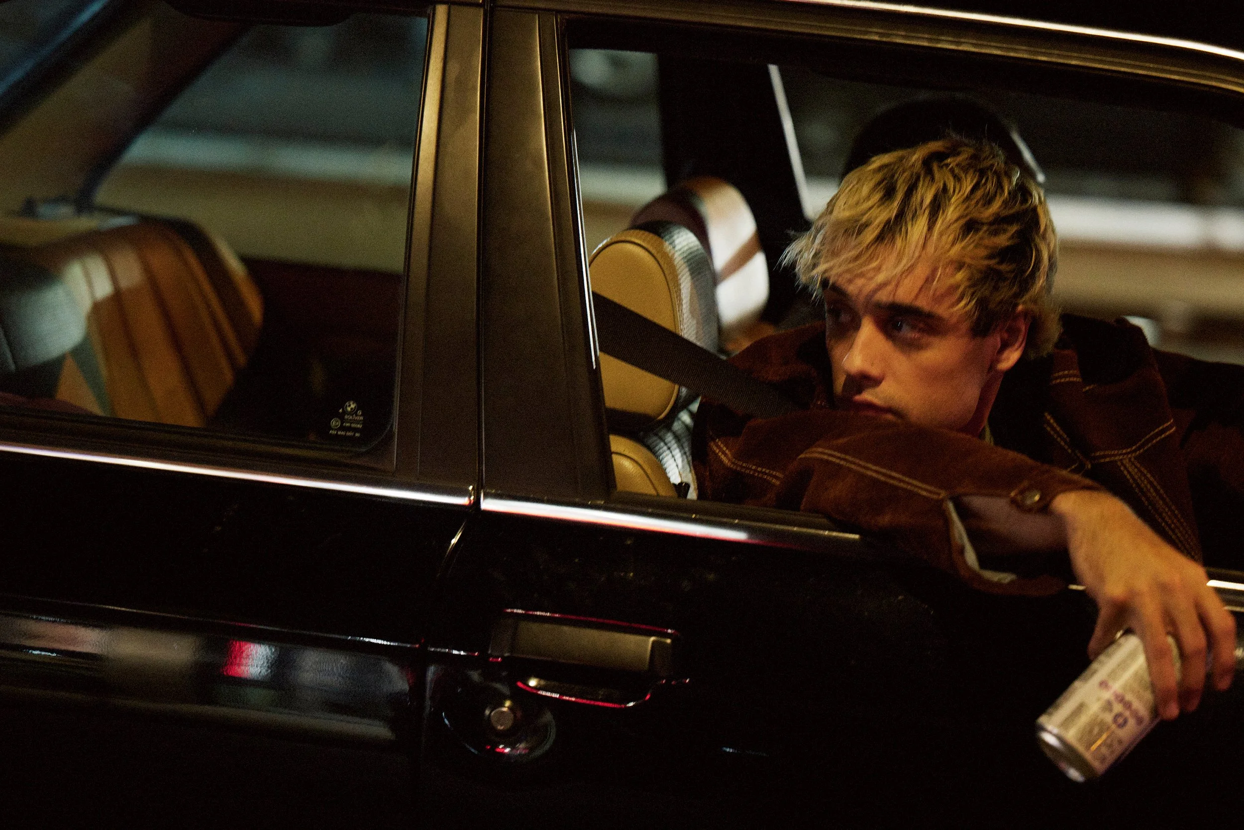A young man with blond hair sitting in the backseat of a black car, looking out the window while holding a can of beer.