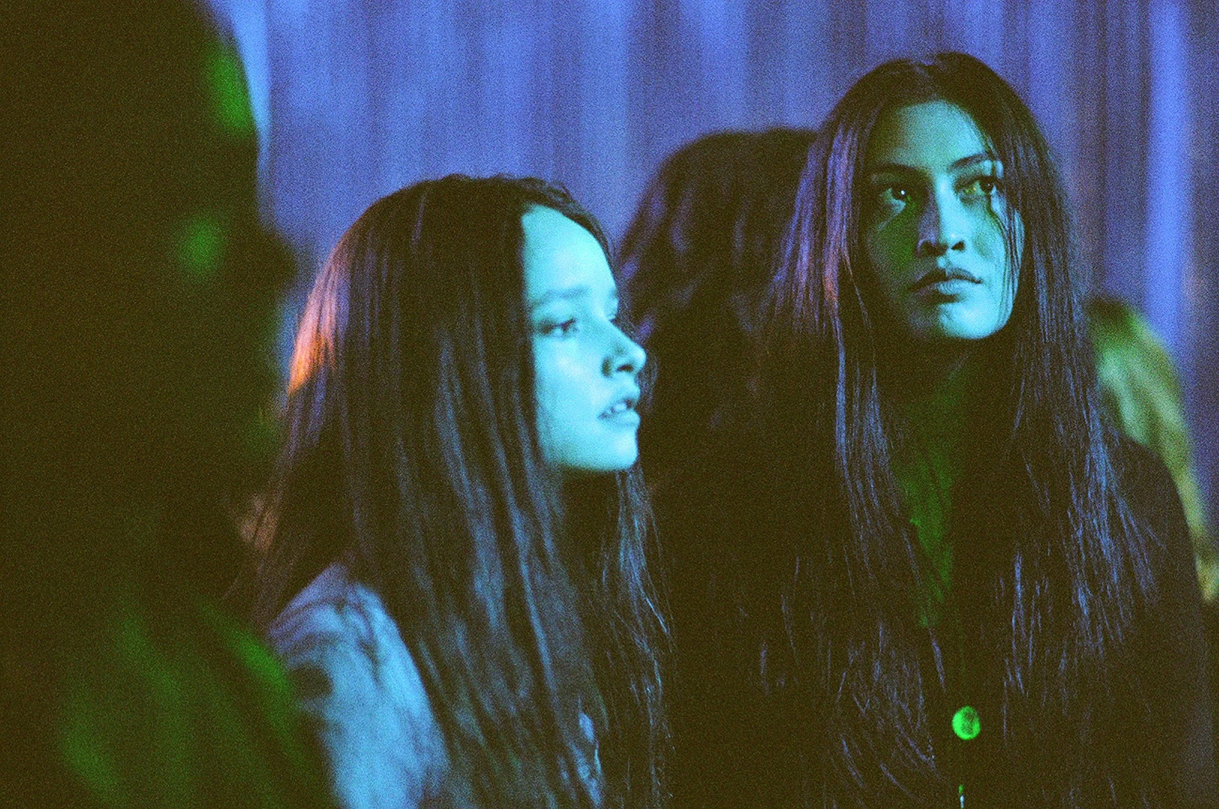 Two women with long, wet hair standing in a dimly lit room with blue and green lighting, looking to the side with serious expressions.