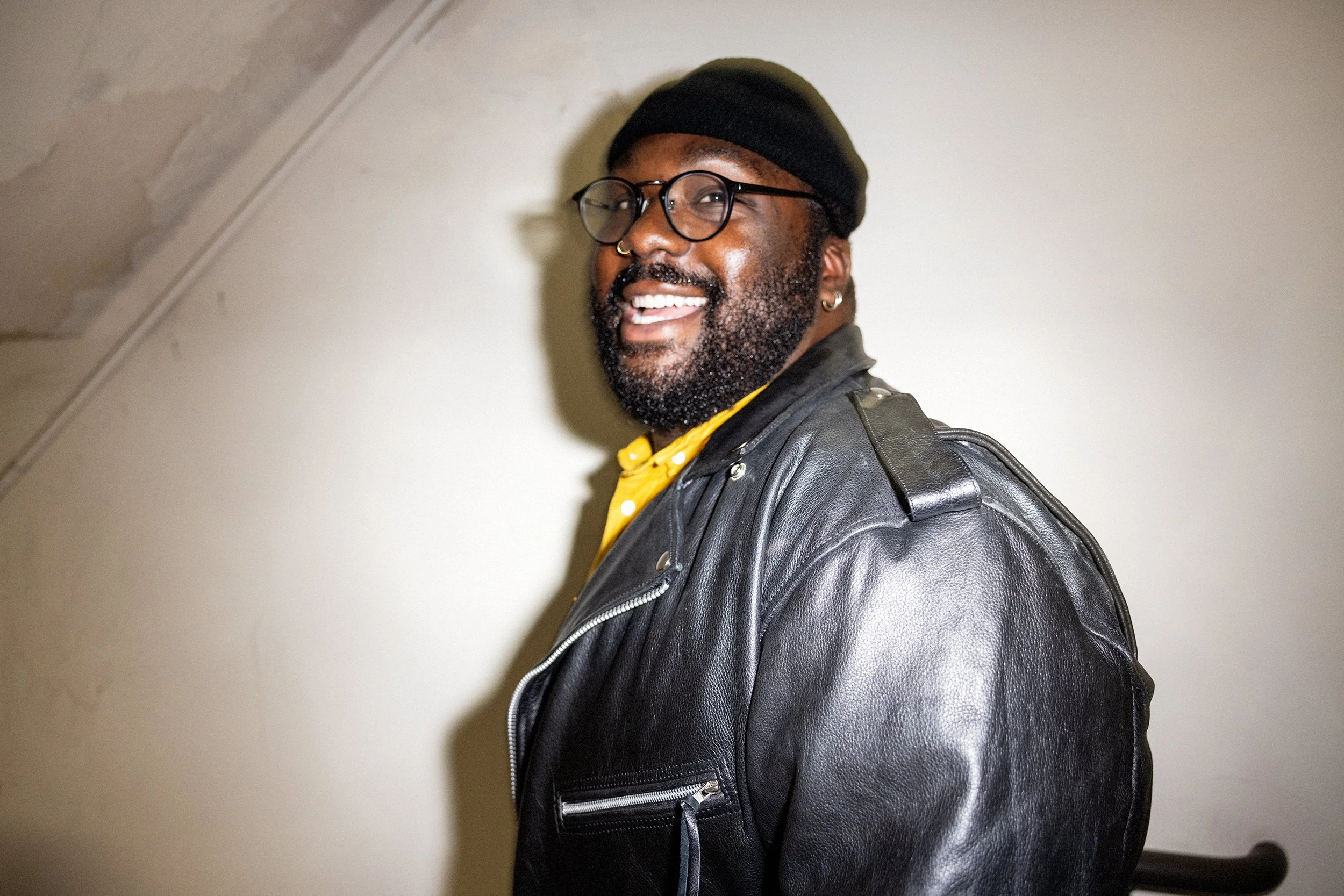 A smiling man with a beard, glasses, and a nose ring, wearing a black beanie, a leather jacket, and a yellow shirt, standing indoors against a plain wall.