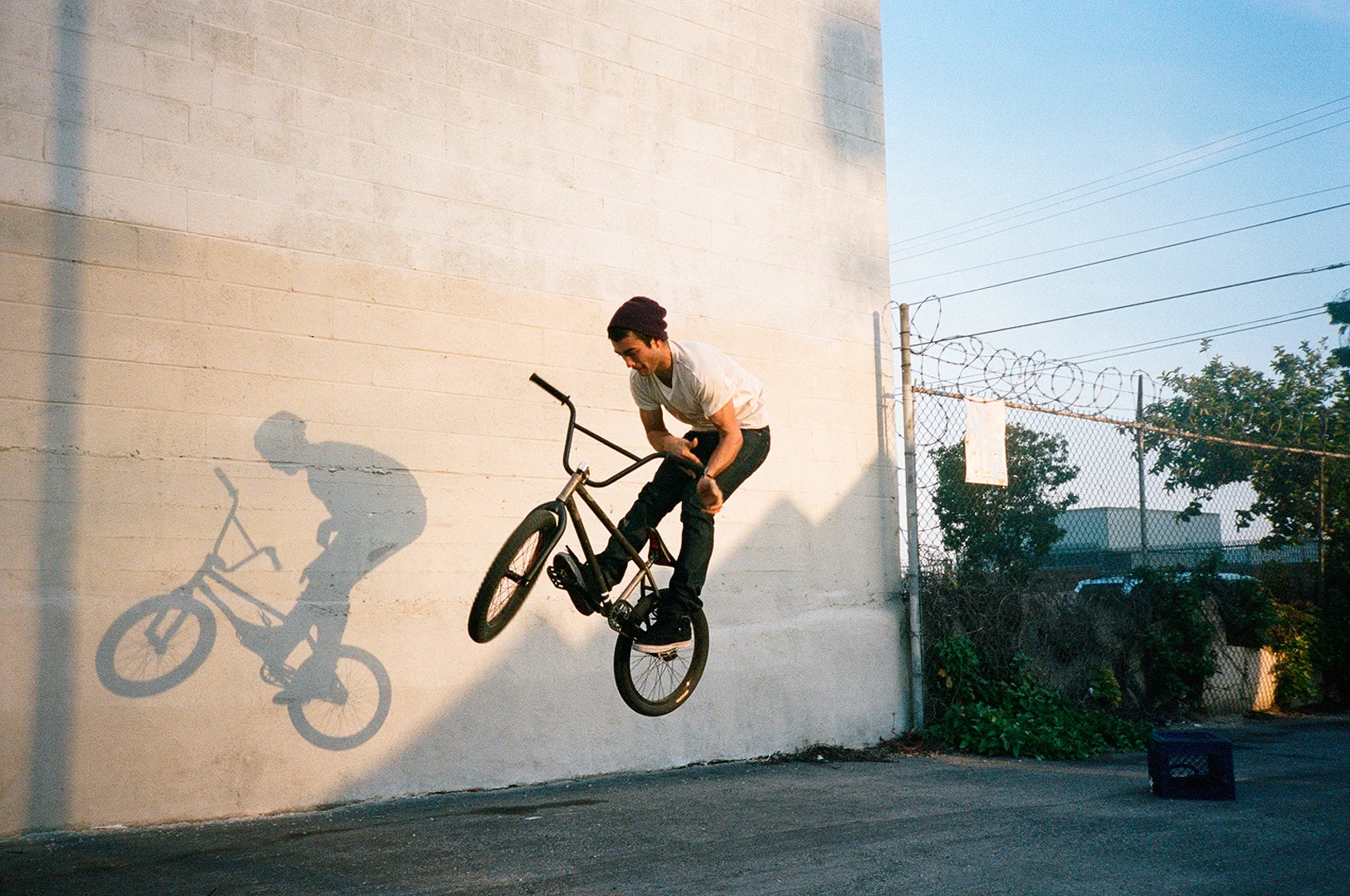 A person in a white t-shirt, black pants, and maroon beanie is performing a BMX trick on a half-pipe against a plain beige wall. The person's shadow is cast on the wall, showing the movement. The scene is outdoors during sunset or sunrise, with a cha