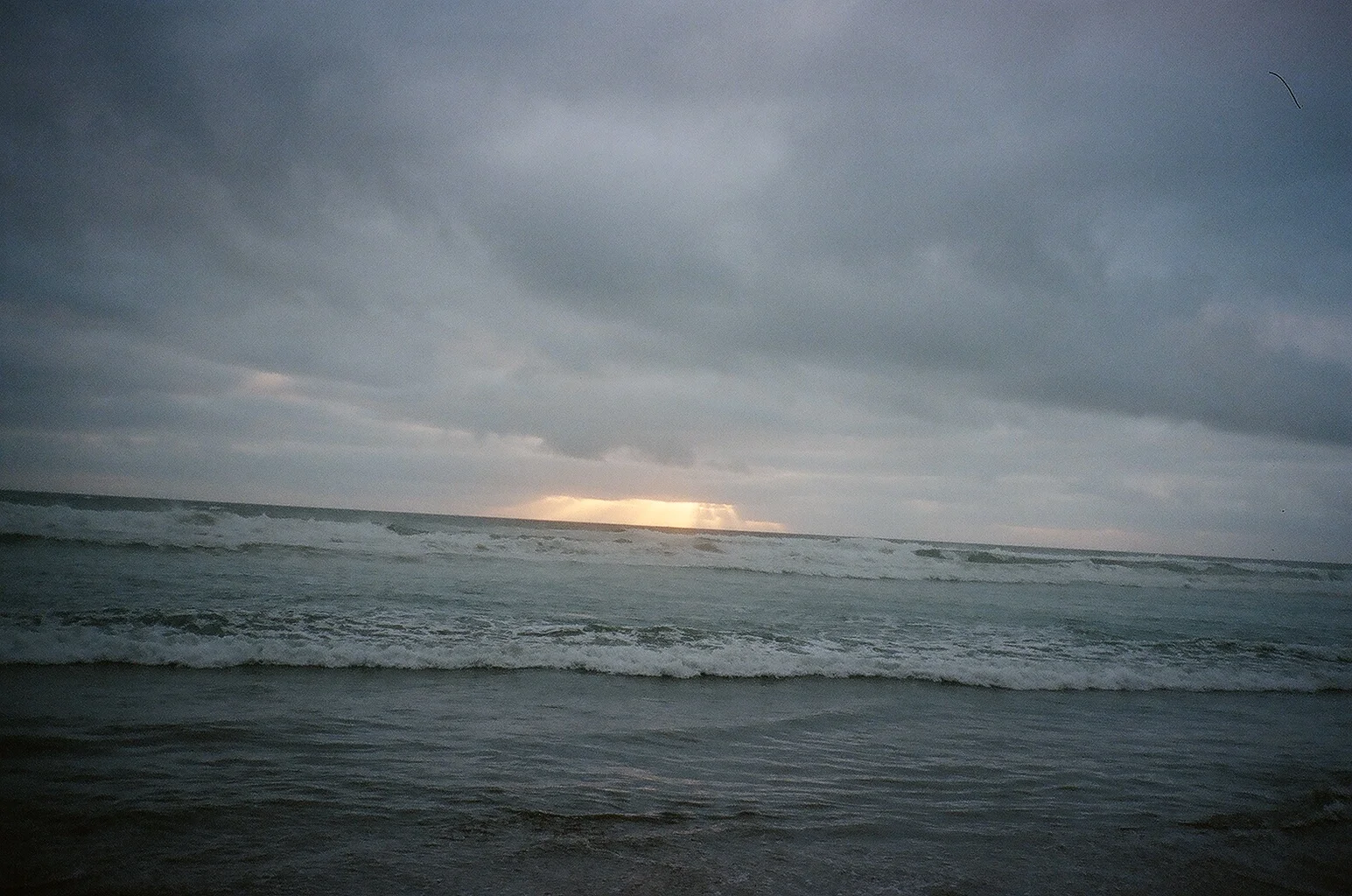 Overcast beach scene with gray clouds, waves crashing onto the shore, and a hint of light near the horizon.