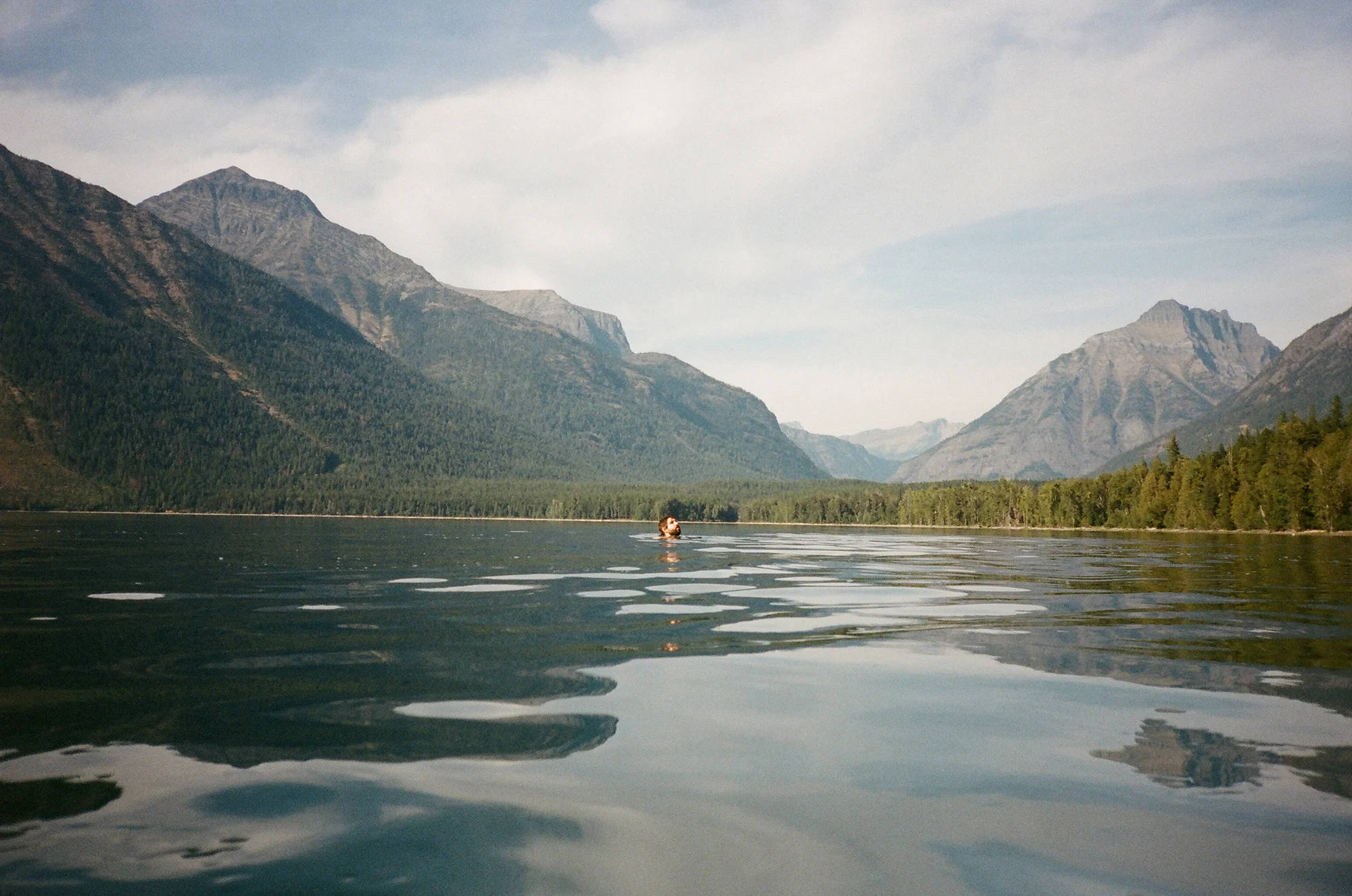 A person swimming in a large, calm lake surrounded by tall mountains with a forested shoreline under a partly cloudy sky.