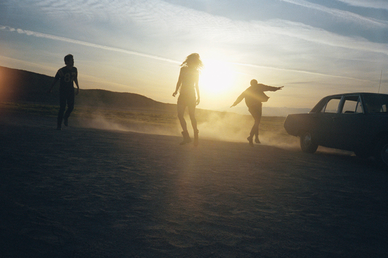 Three people dancing on a desert landscape at sunset near an old car, with dust clouds and mountains in the background.