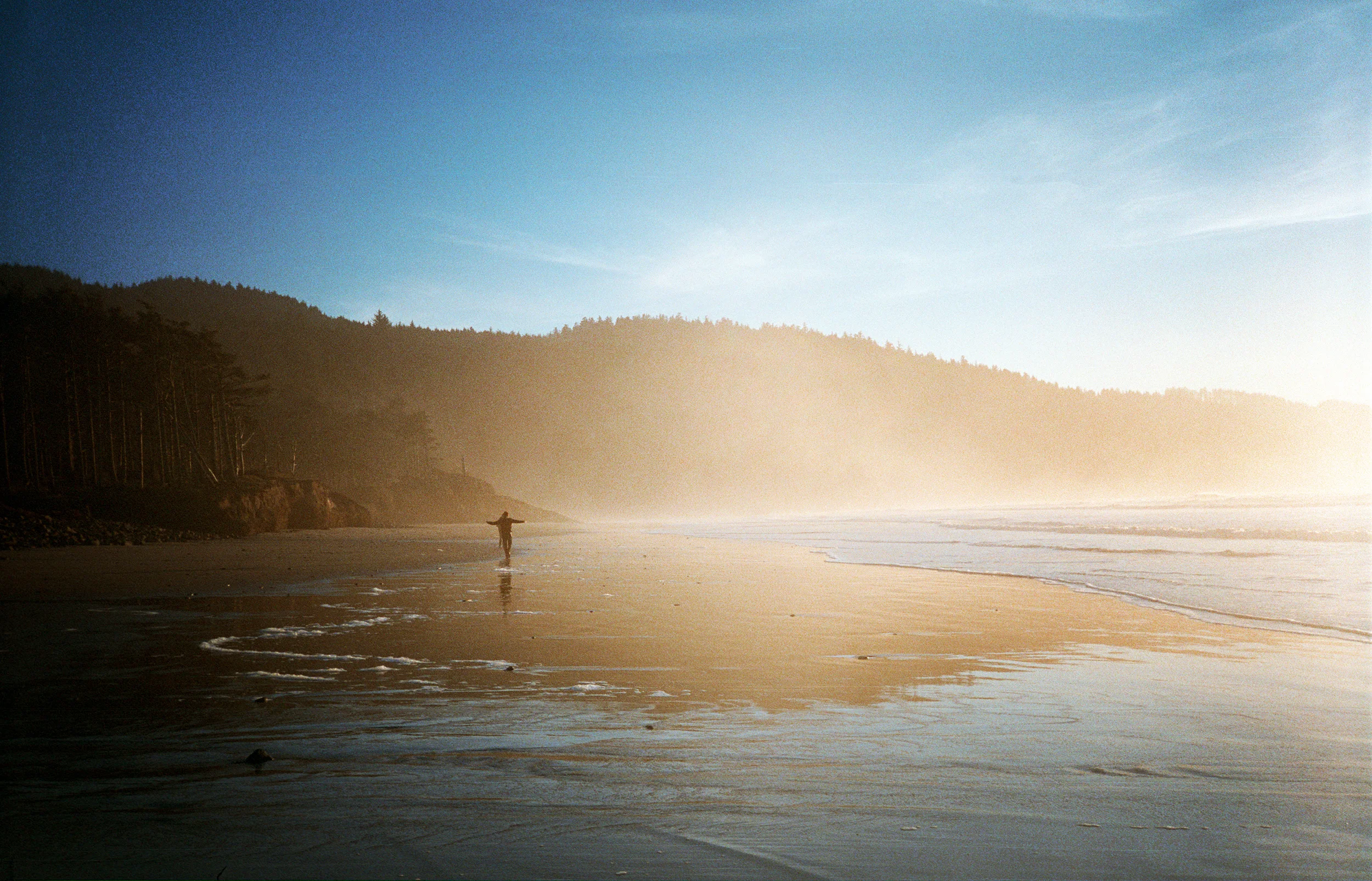 A person walking on a sandy beach toward the ocean with mist and hills in the background during sunset or sunrise.