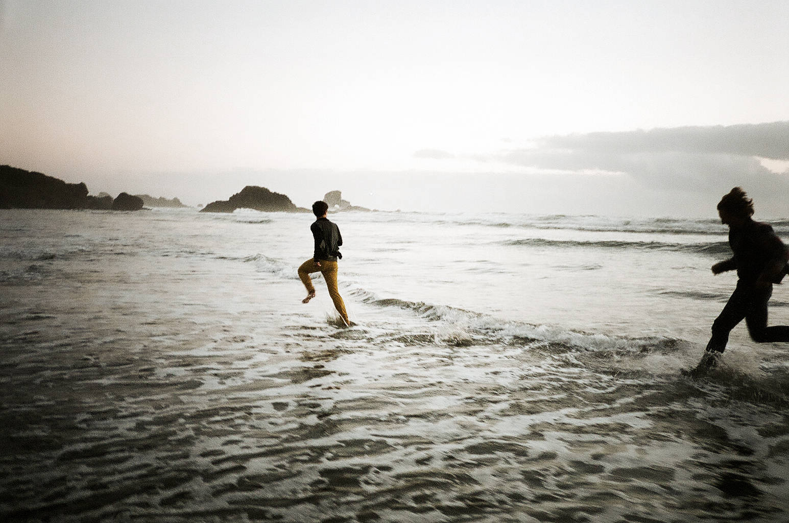 Two people running in shallow ocean waters near a rocky shoreline during sunset or sunrise.