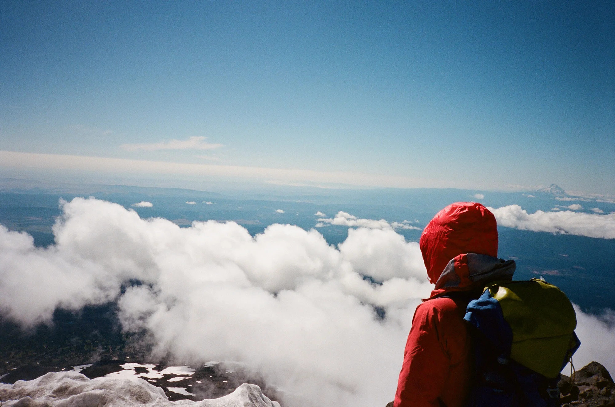 Person in a red jacket and backpack sitting on a snow-covered mountain summit, overlooking clouds, with a blue sky and distant mountain peaks in the background.