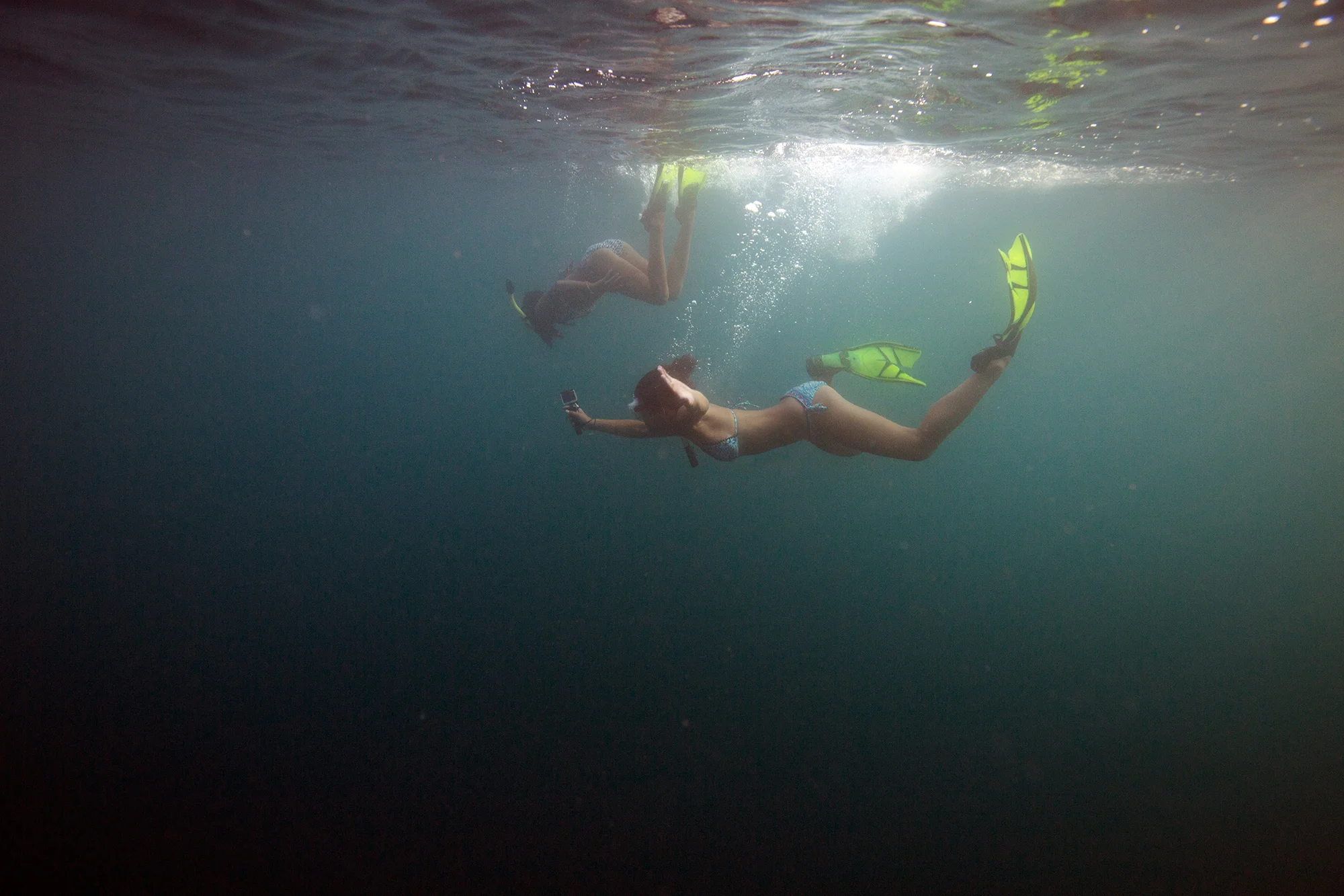 Two women are swimming underwater with one holding a phone and wearing a bikini, the other wearing a bikini and looking downward.