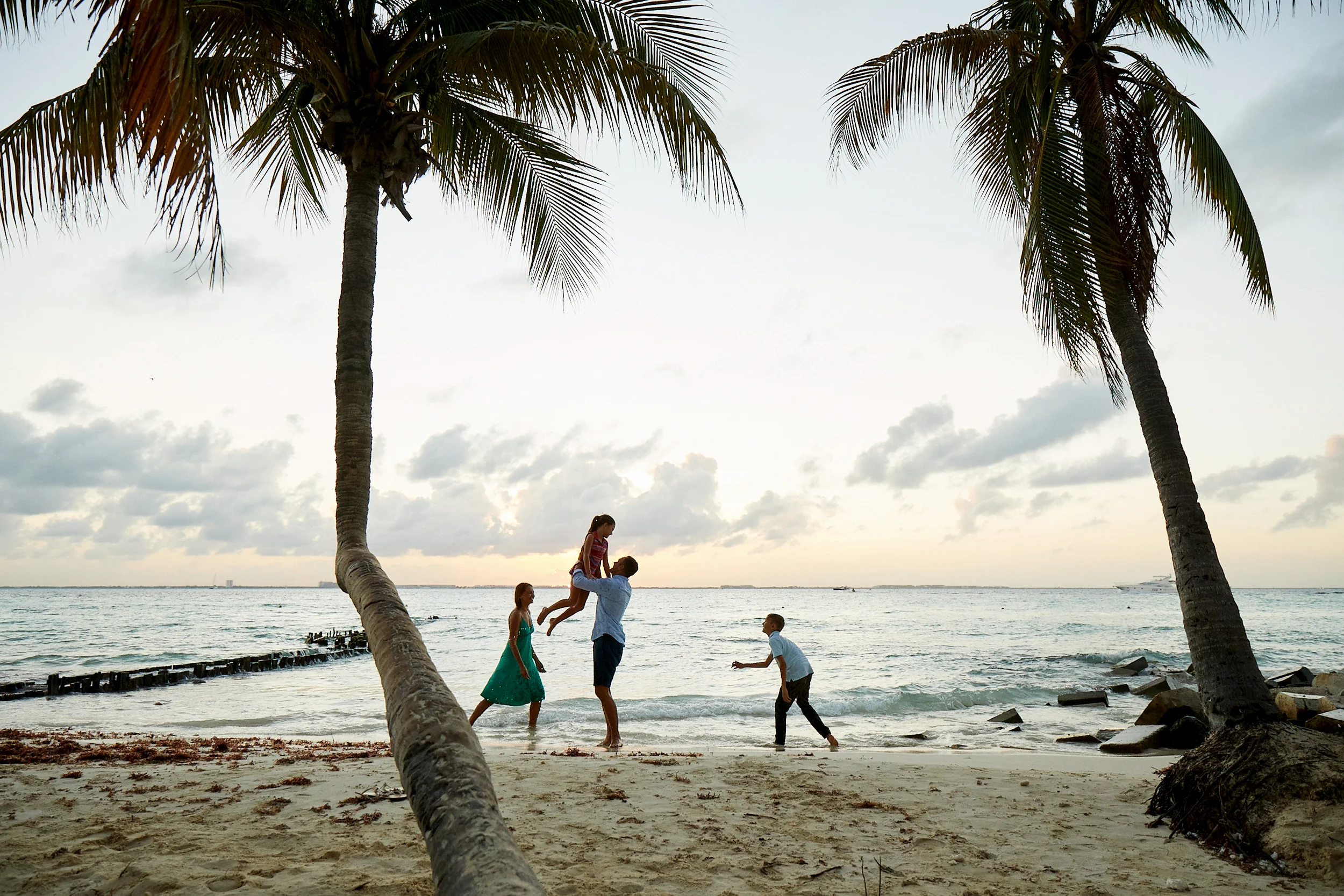Family playing and having fun on a beach at sunset, with palm trees and the ocean in the background.