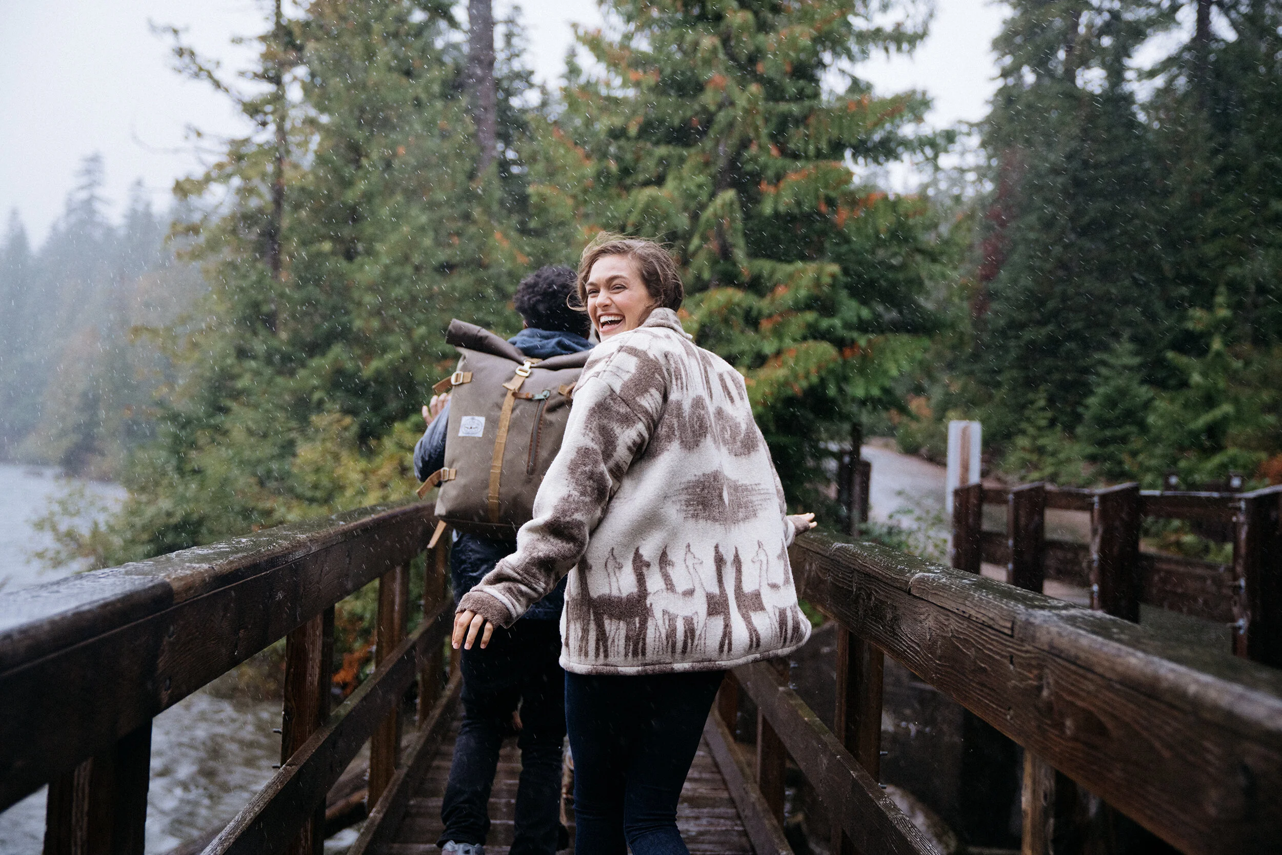 People smiling and walking on a wooden bridge in a forest during rain, with trees and a lake in the background.
