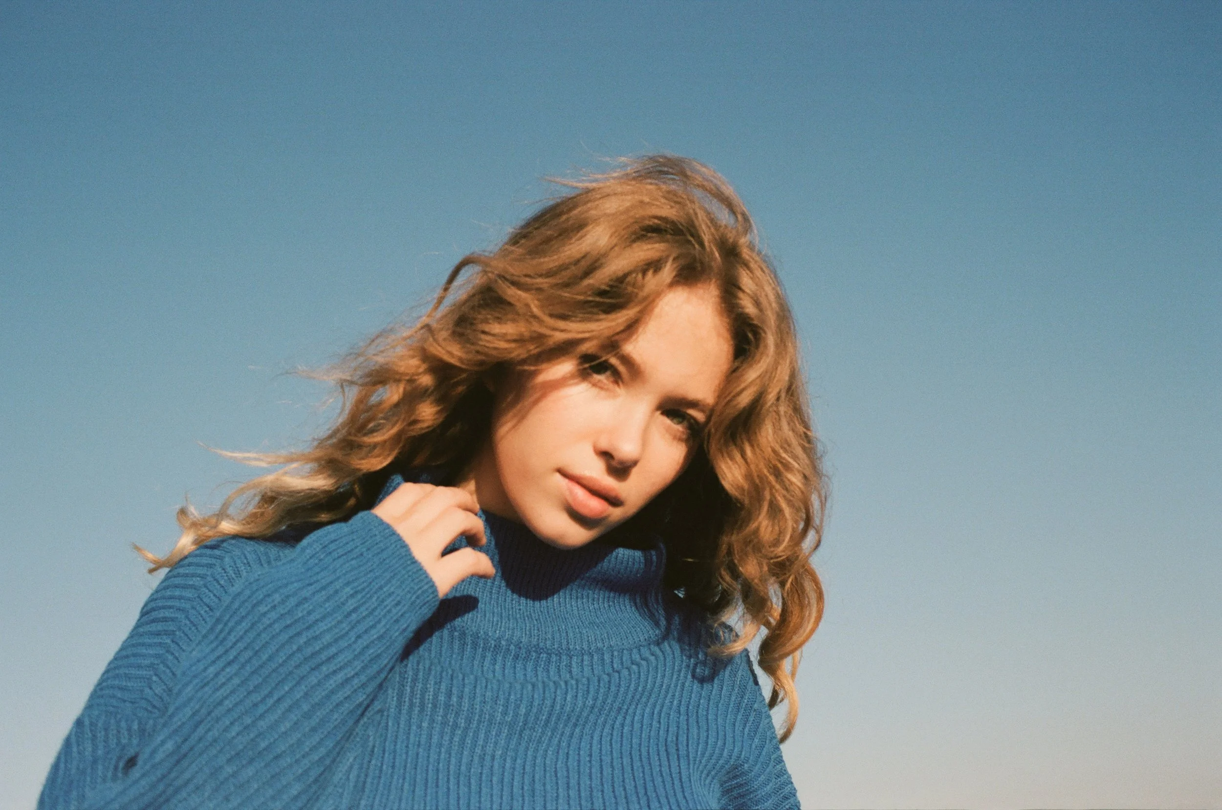 A young woman in a blue sweater standing outdoors against a clear blue sky, with curly hair and a slight smile.