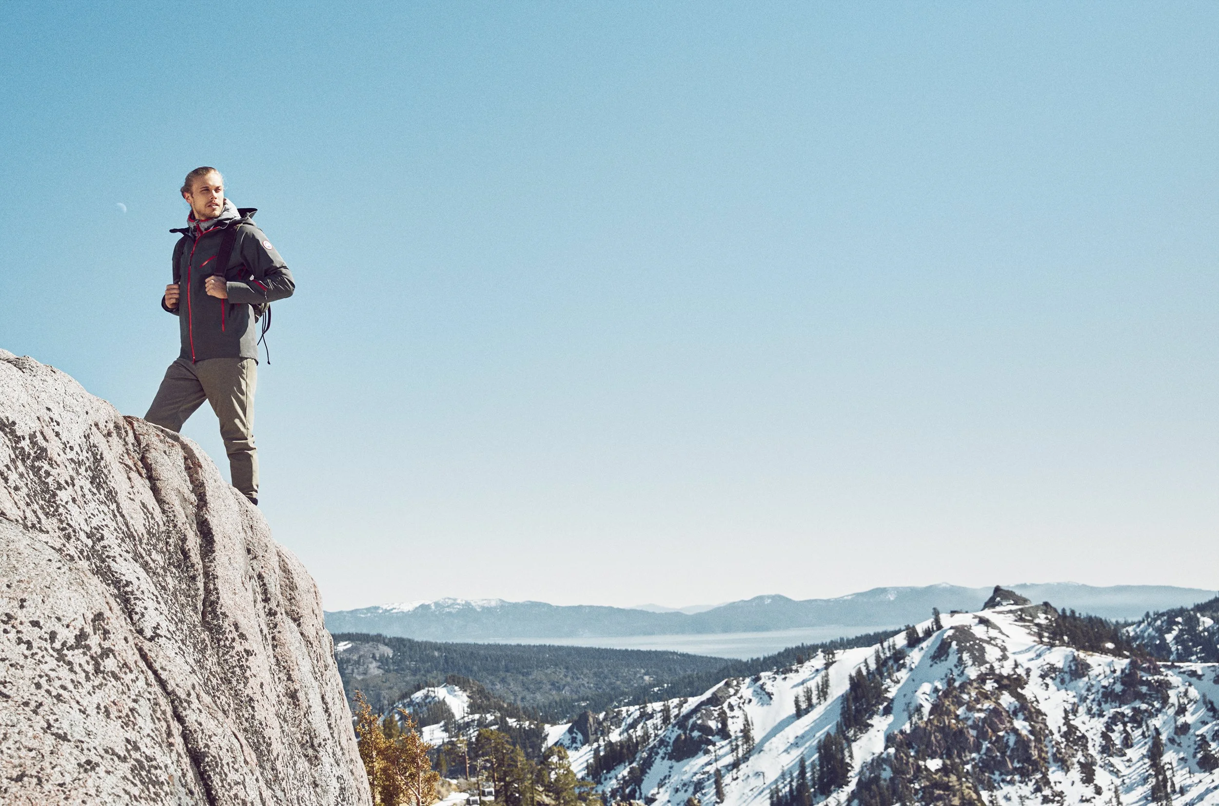 A man standing on a rocky mountain ledge, overlooking snow-covered mountains and a clear blue sky.