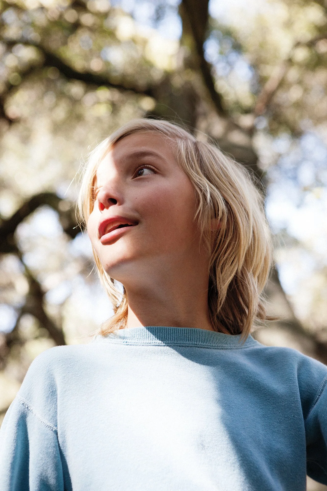 A young woman with blonde hair looking up outdoors in a natural setting with trees and sunlight.