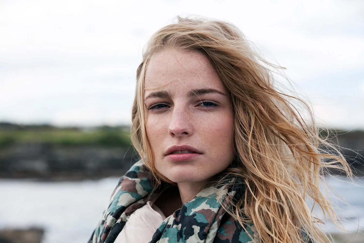 Young woman with wavy blonde hair outdoors, wearing a camouflage jacket, looking into the distance with a serious expression.