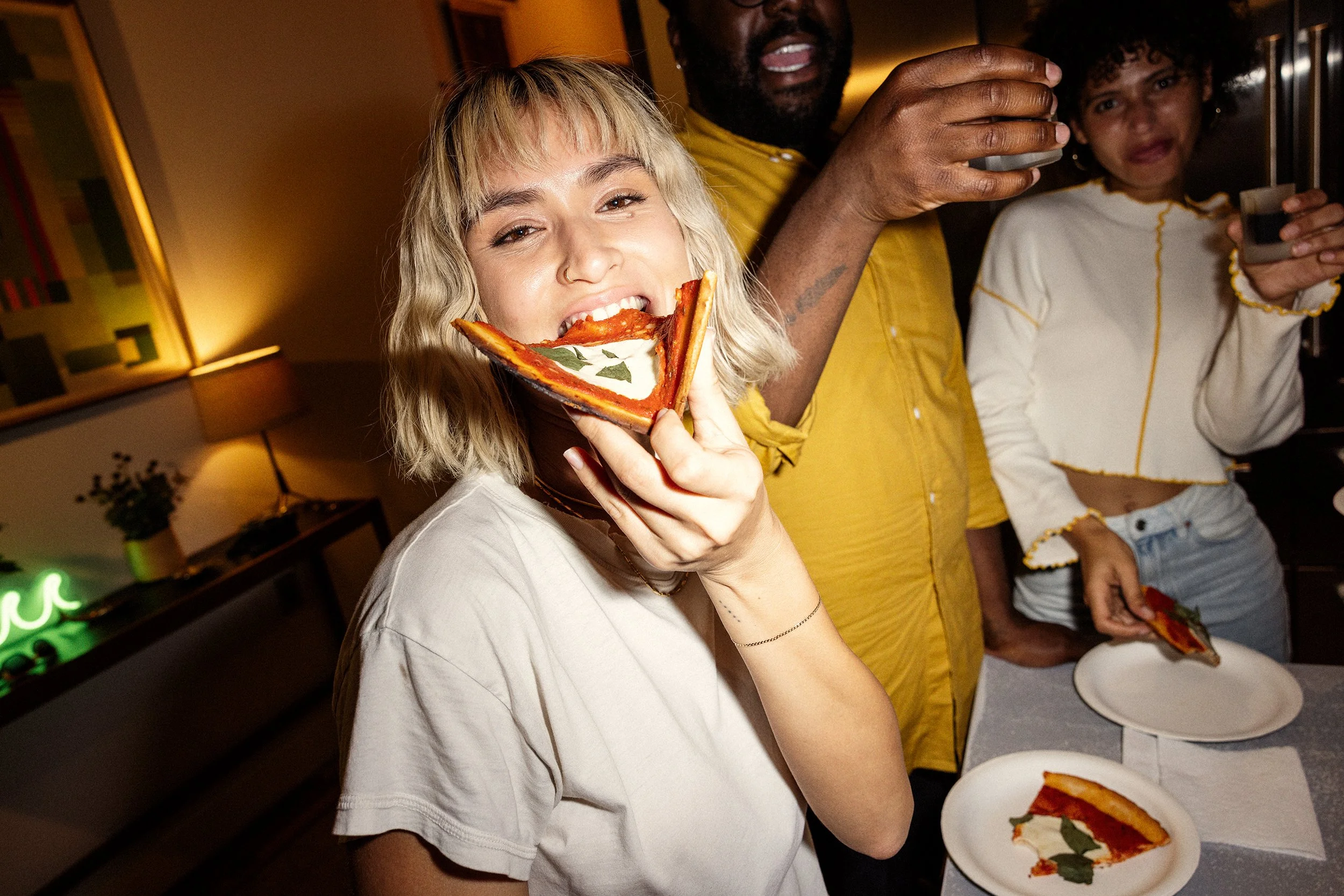 A young woman with blonde hair and a nose ring eating a slice of pizza. Other people are in the background, one person holding a drink and another holding a phone, at a social gathering.