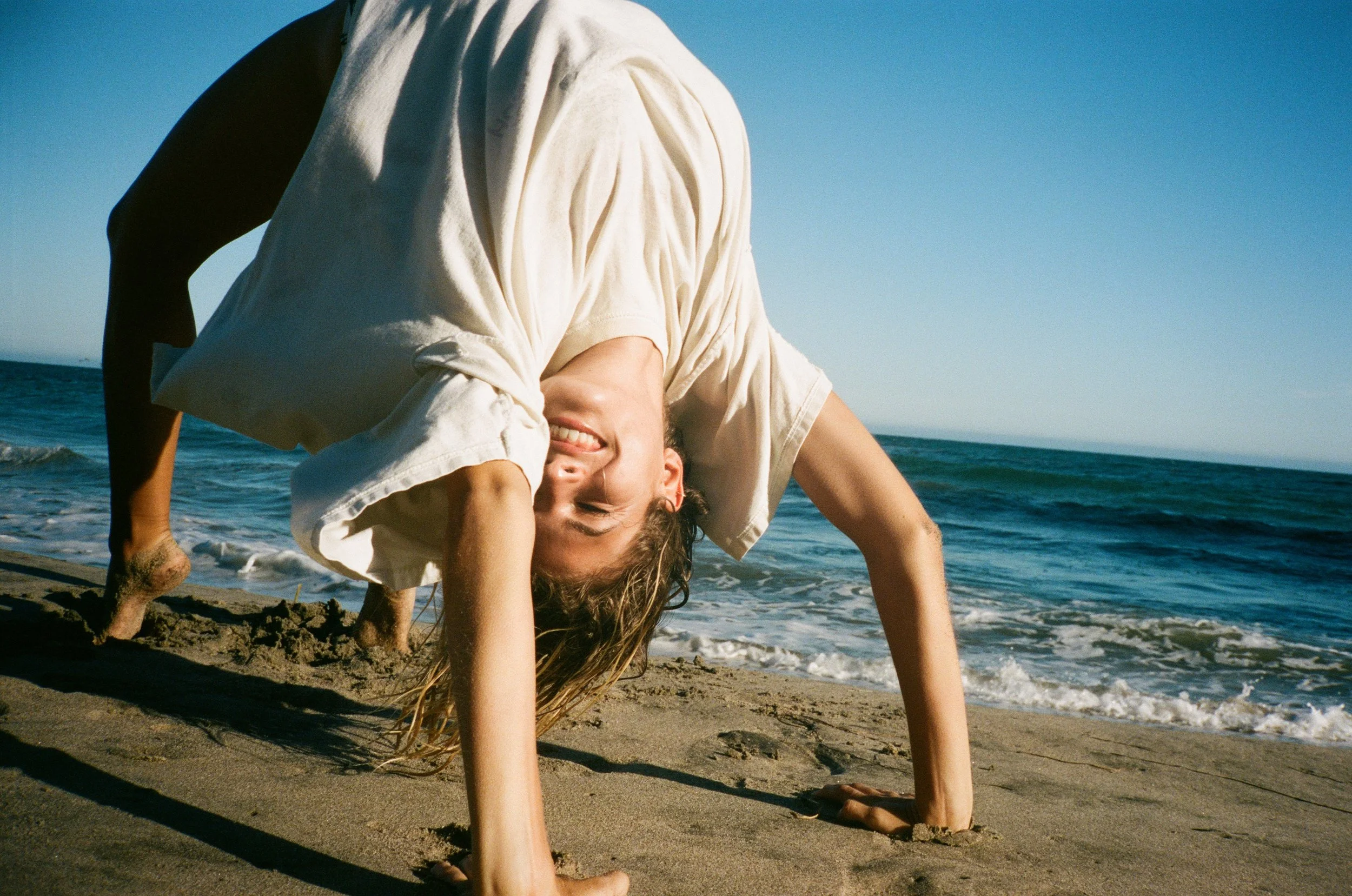 Person in a white t-shirt performing a backbend on a sandy beach with the ocean in the background, smiles while looking at the camera.