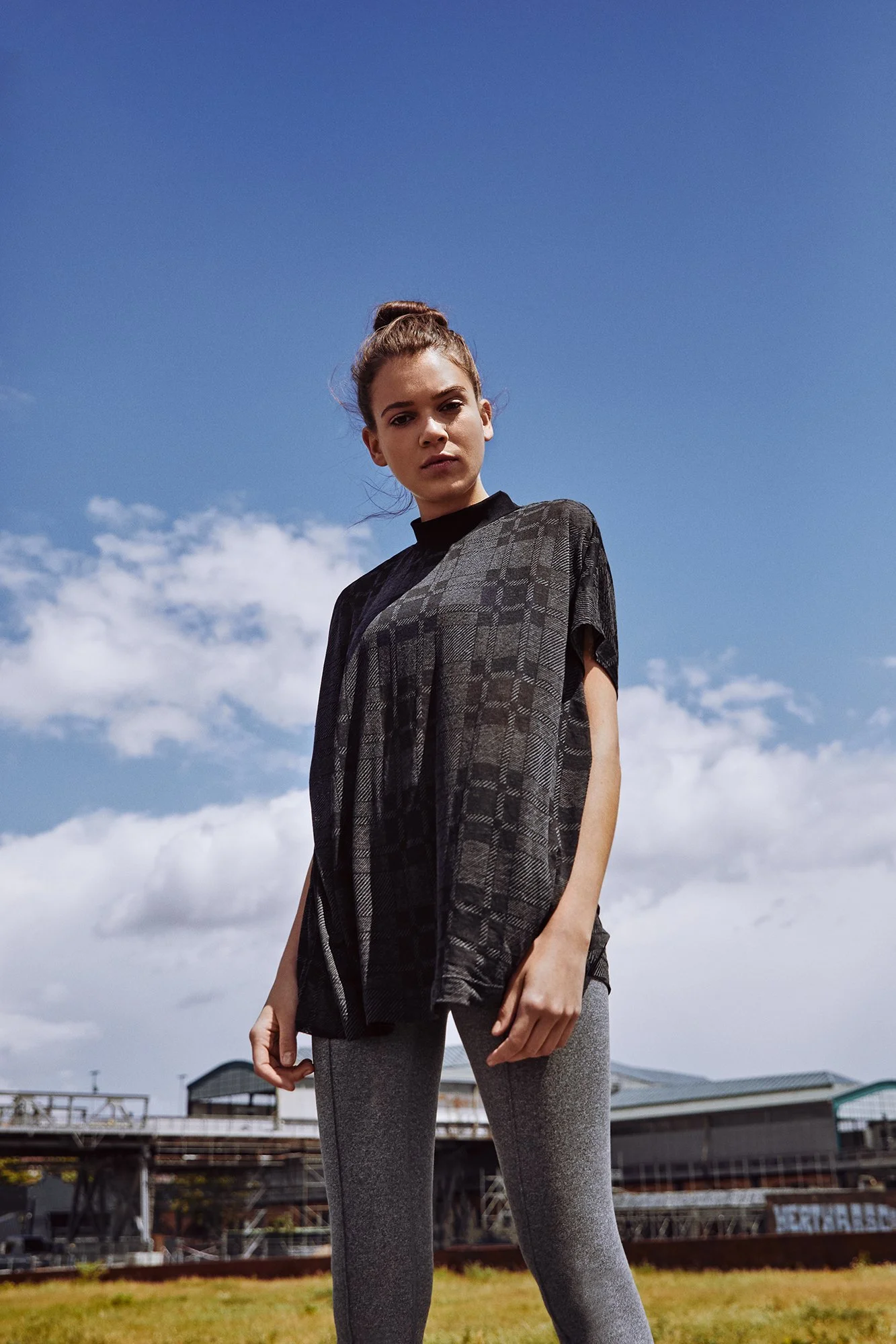A woman standing outdoors against a blue sky with clouds, wearing a black and gray patterned top and gray leggings.