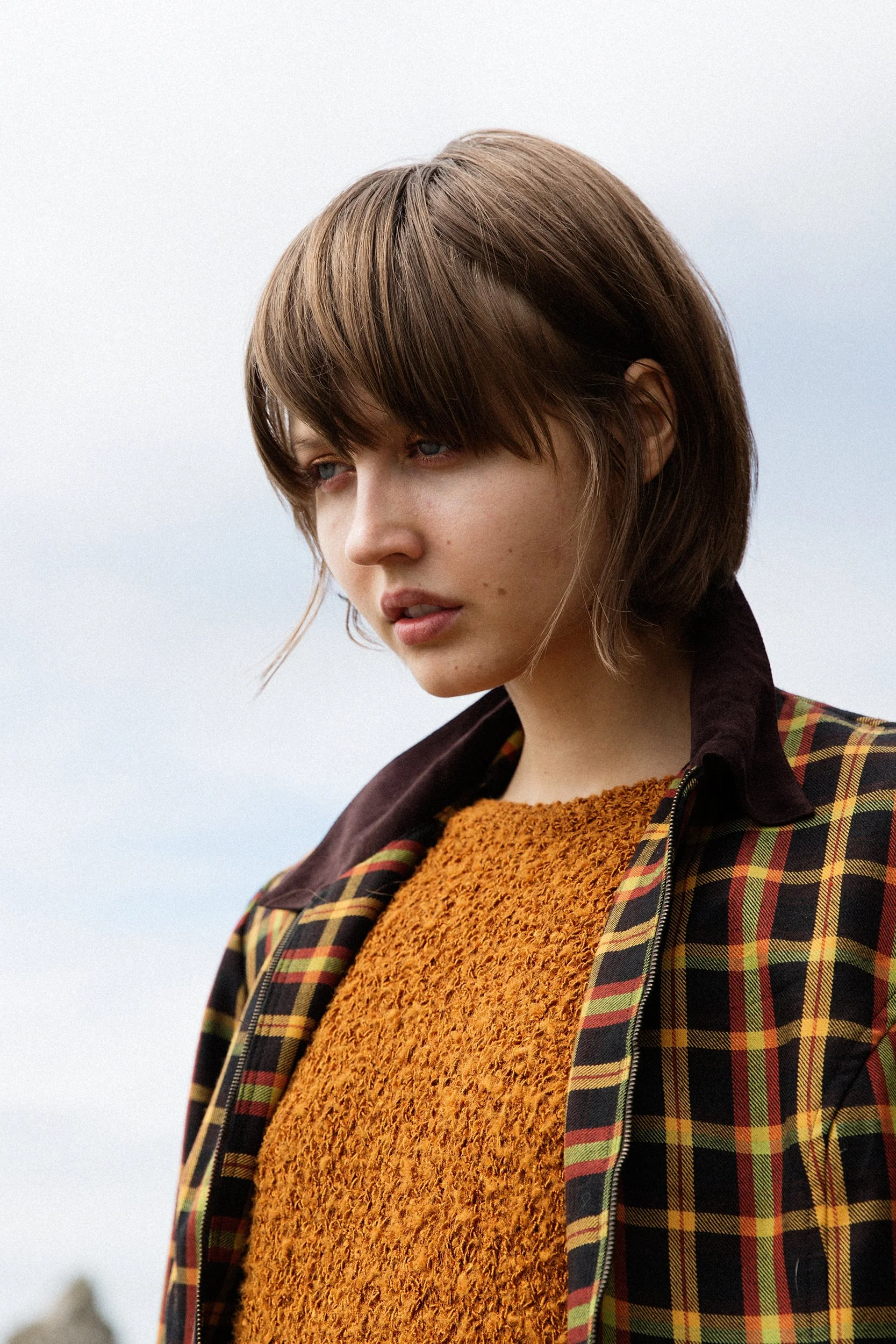 Close-up of a young woman with short brown hair wearing a yellow textured sweater and a plaid jacket, outdoors against a cloudy sky.