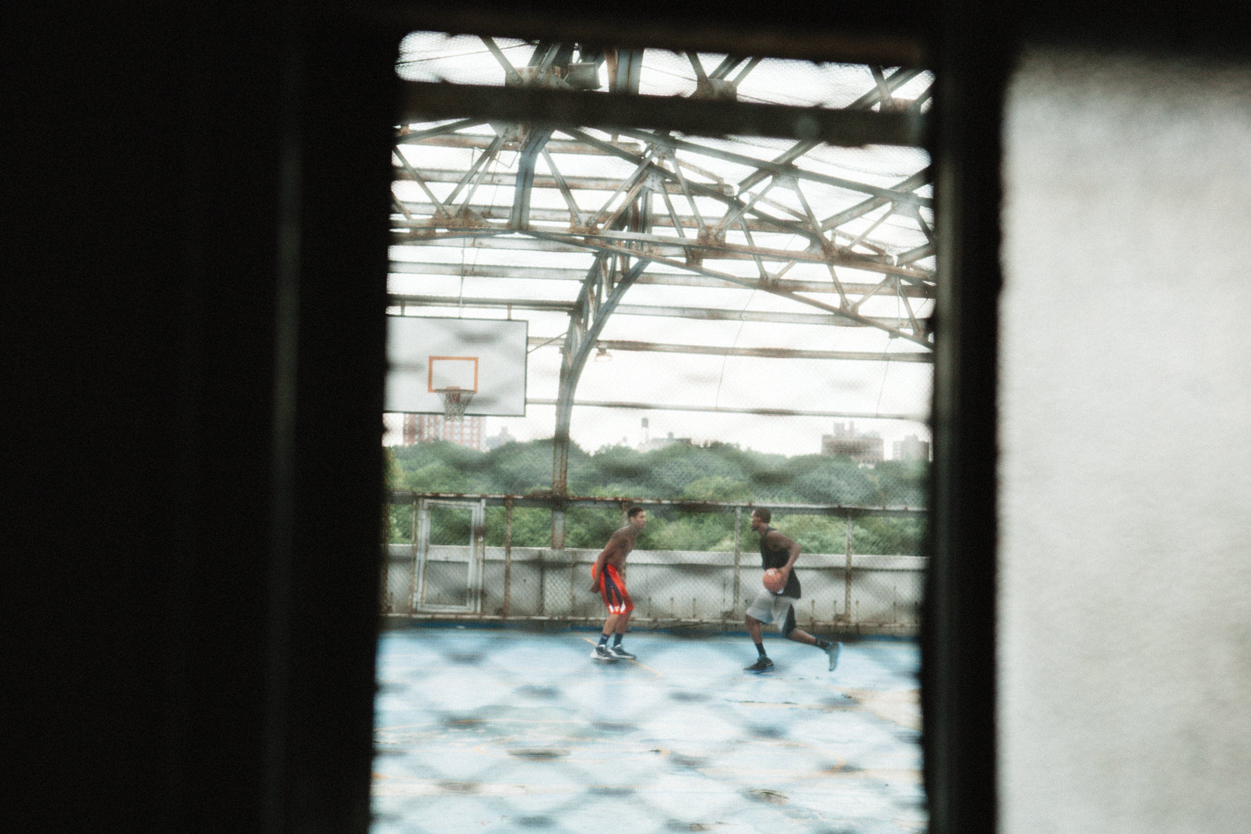 Two people playing basketball on an outdoor court, viewed through a window with a wire mesh. The court has a blue surface, and there is a basketball hoop in the background. The scene is set under a large, rusty metal shelter, with city buildings and 