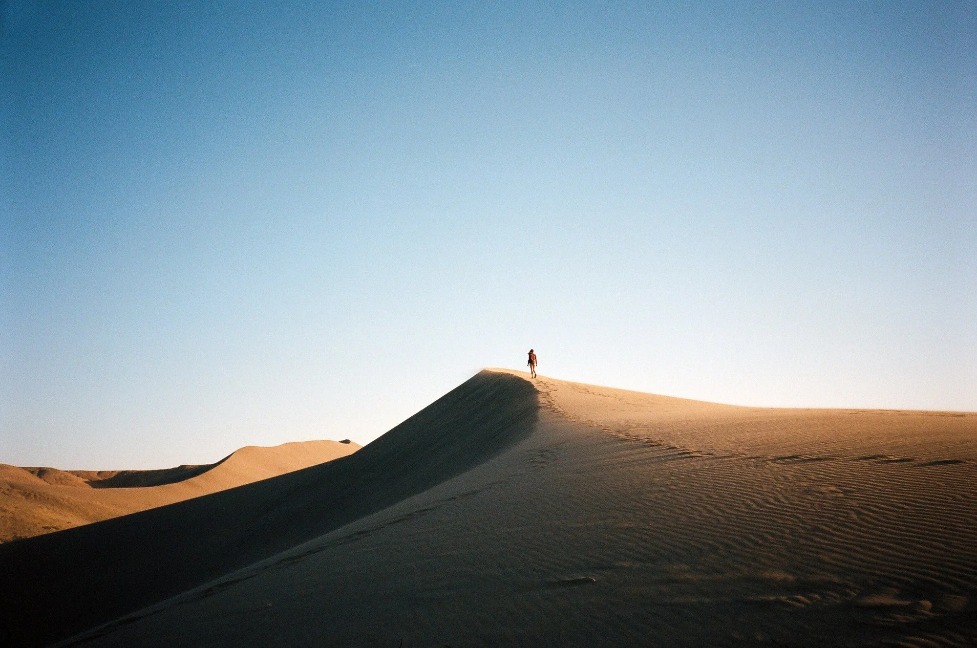 A person walking on top of a sand dune in a desert with a clear blue sky.