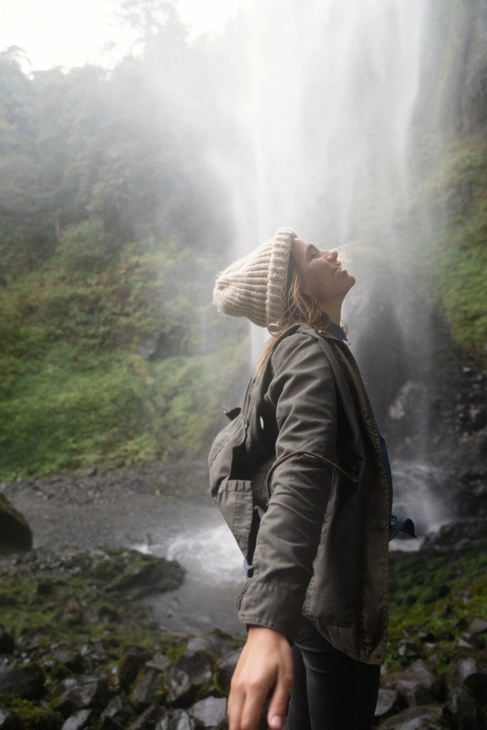 A woman in a beige knit beanie and green jacket standing in front of a waterfall with mist and greenery.