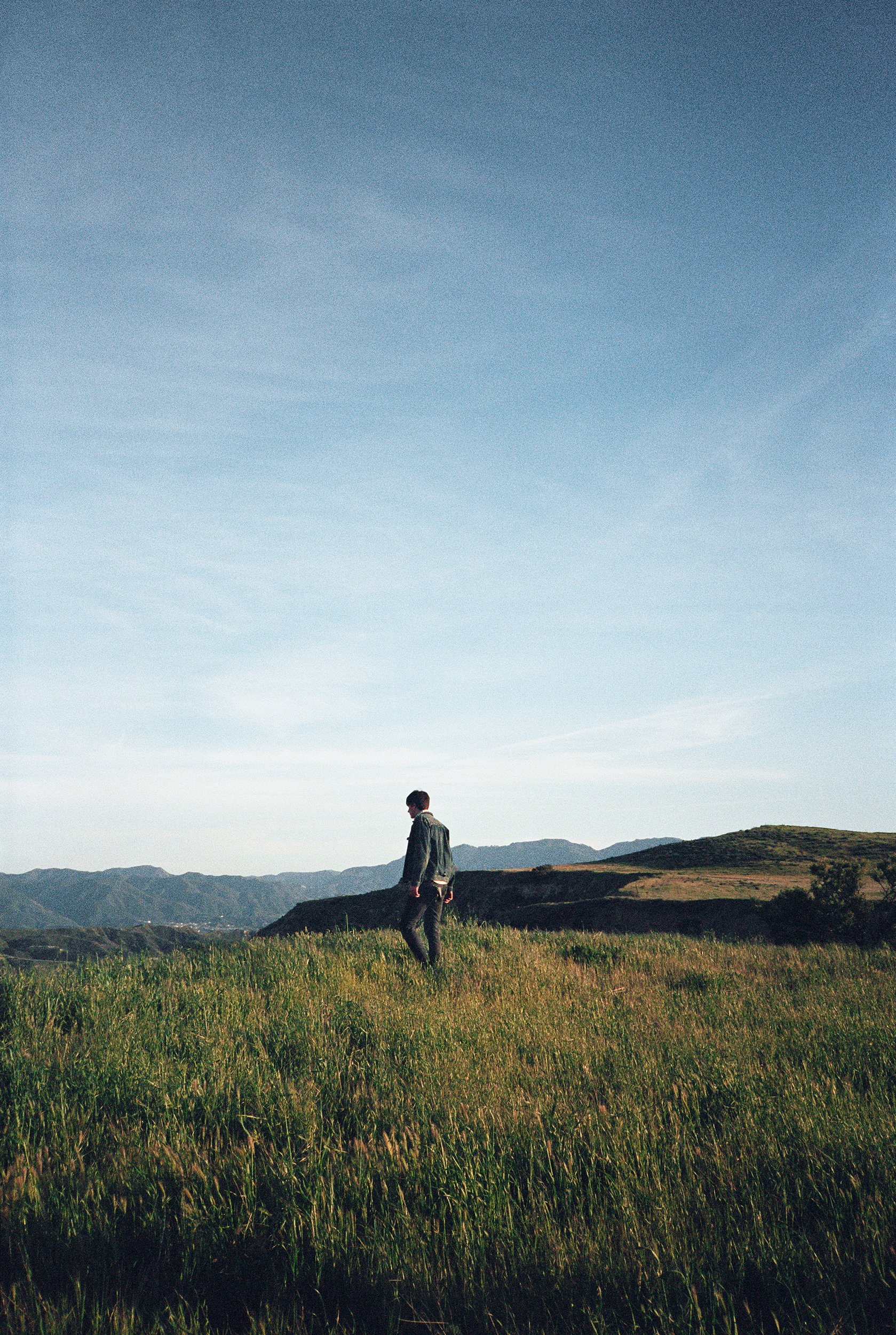 A person walking through tall grass in a field with mountains in the distance under a clear blue sky.