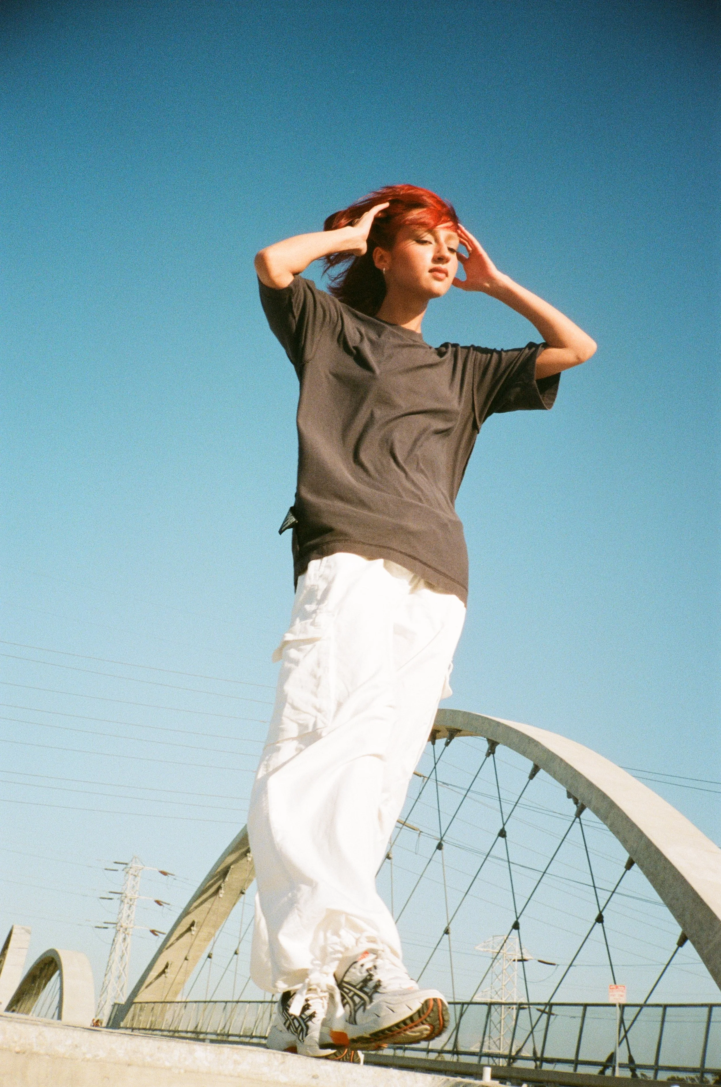 A young woman with red hair stands on a bridge with her hands on her head, gazing into the distance against a clear blue sky.