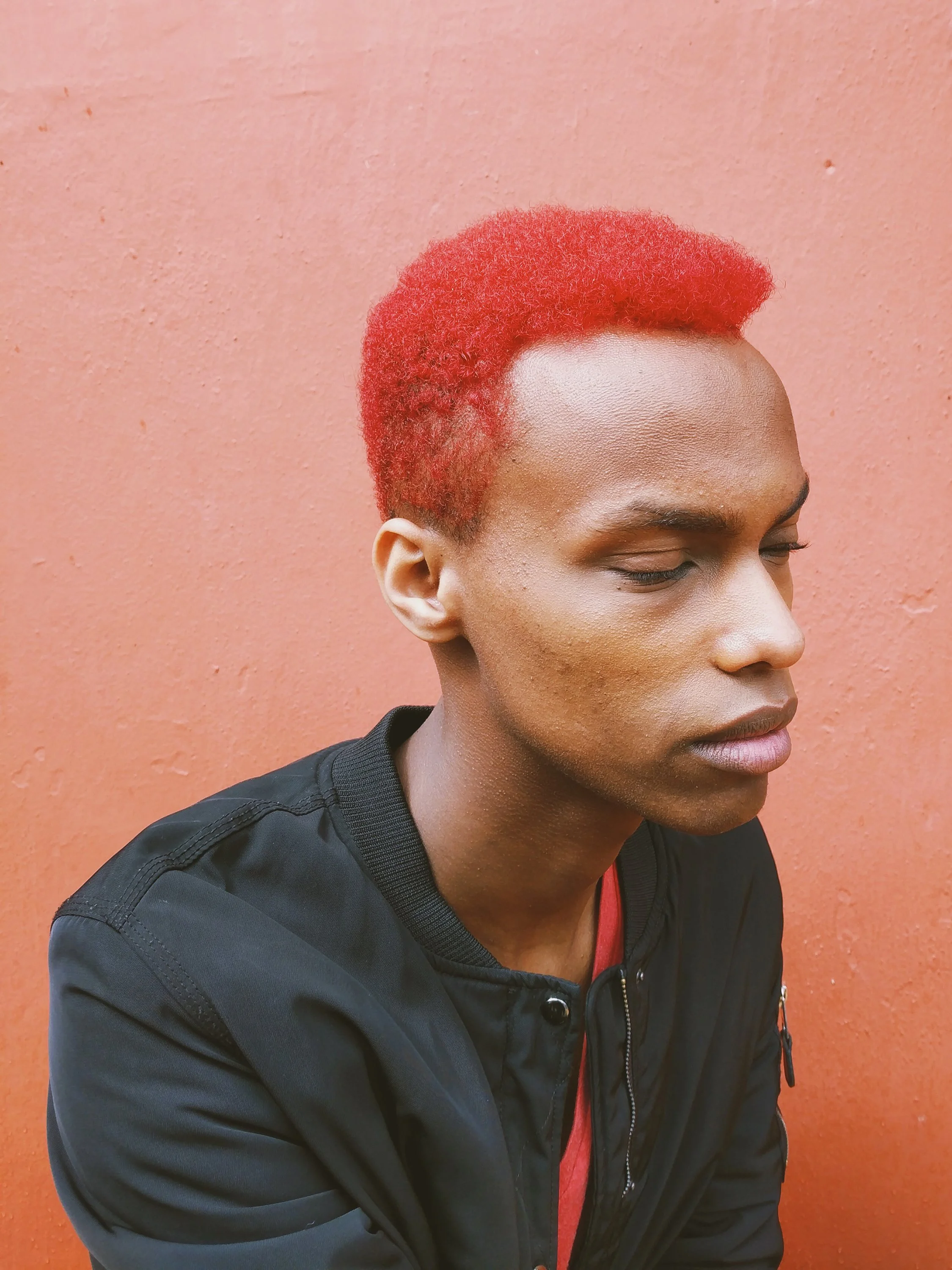 Portrait of a person with vibrant red hair, closed eyes, and wearing a black jacket against a solid pink background.