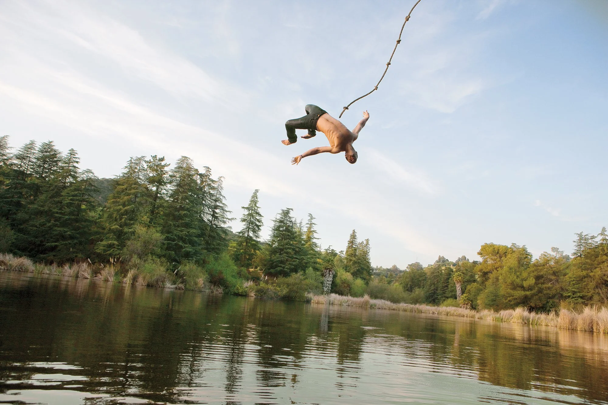 A person sliding on a rope swing over a river, performing a flip in mid-air with trees in the background.