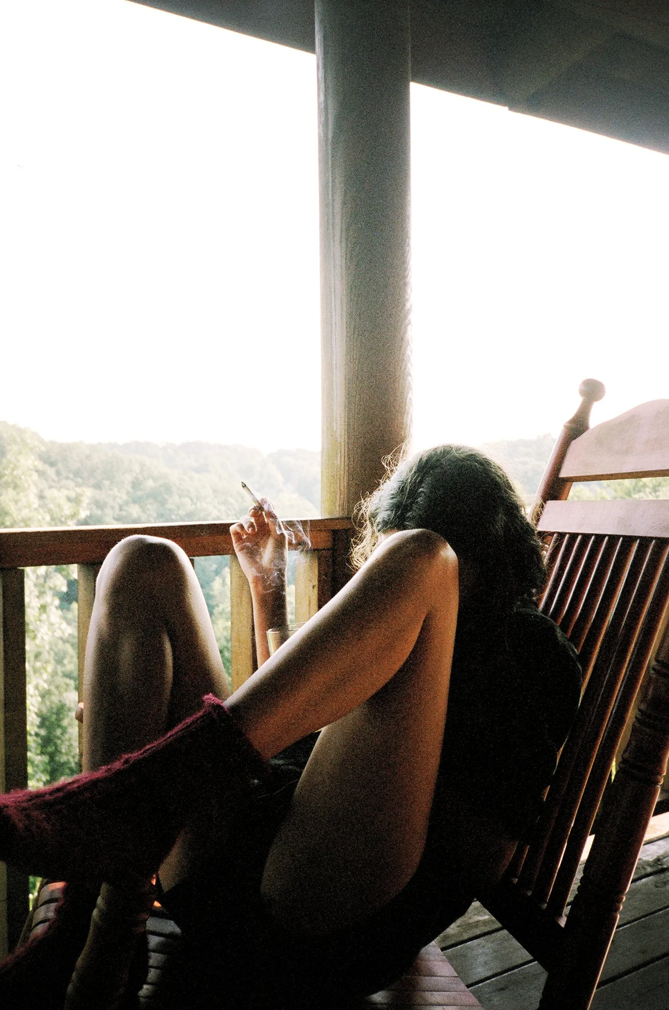 A person sitting on a porch or balcony with a view of trees, smoking a cigarette or joint, with their head resting against a wooden rocking chair.