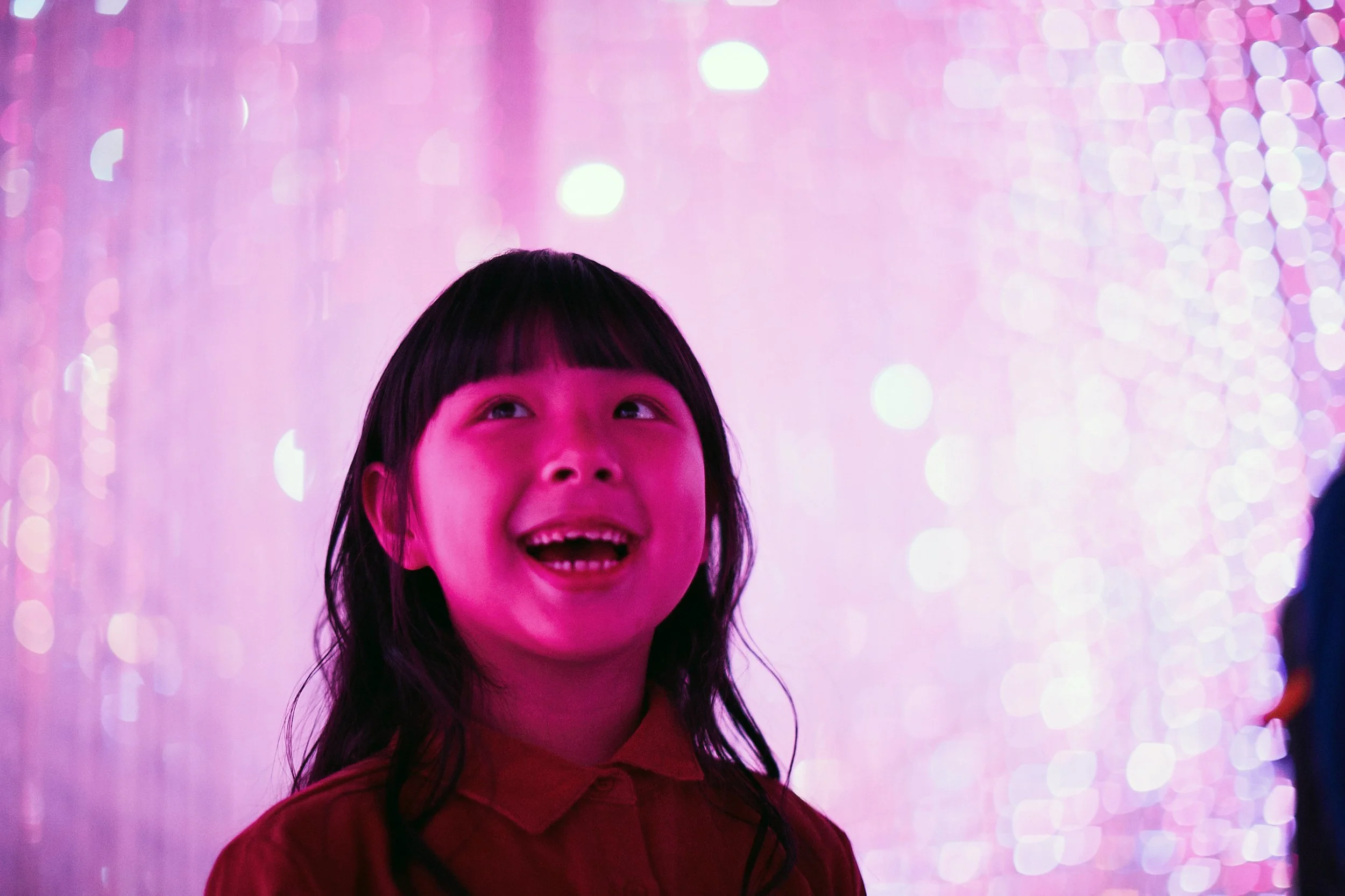 A young girl with dark hair and bangs, smiling and looking upwards, illuminated by pink and purple lights with bokeh effects in the background.