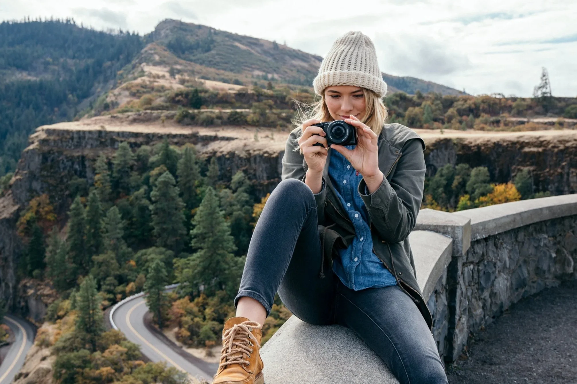 A young woman sitting on a stone ledge in front of a scenic mountainous landscape, holding a camera and looking at it. She is wearing a beige knit hat, gray jacket, blue shirt, black jeans, and tan boots. The background shows winding roads, trees, an