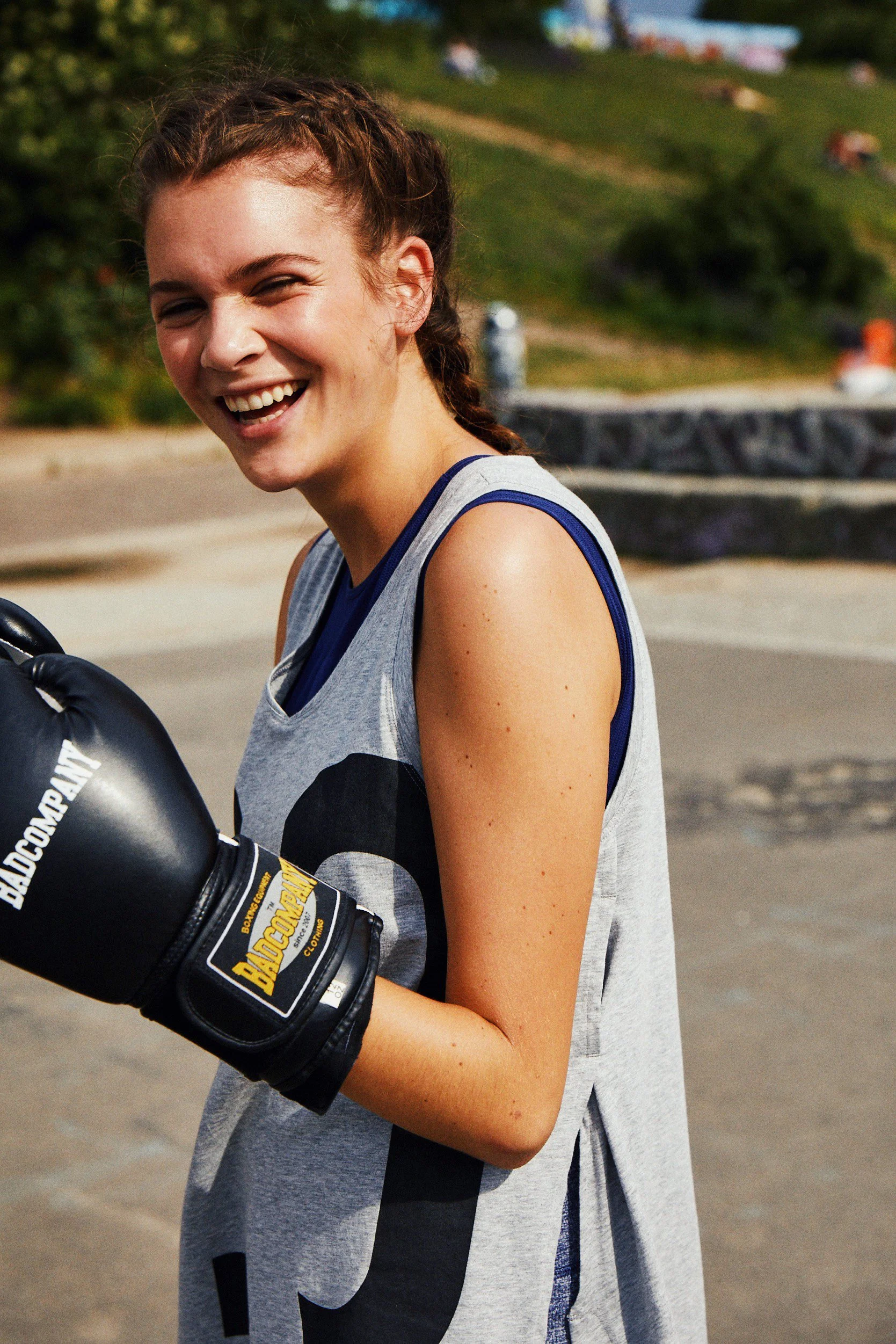Young woman smiling outdoors while holding boxing gloves, dressed in athletic attire.
