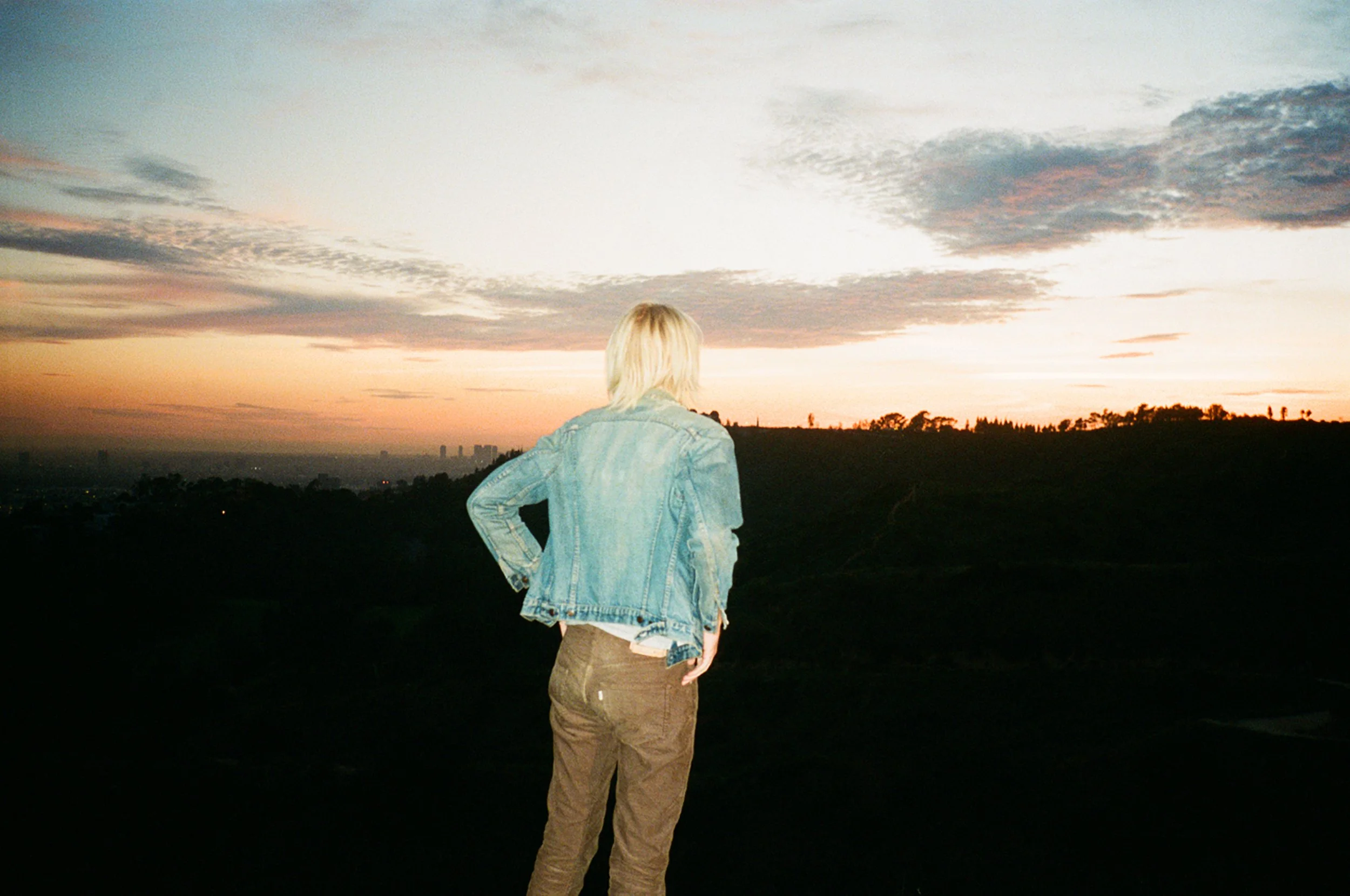 A person with blonde hair wearing a denim jacket and brown pants stands on a hill, looking at a sunset over a landscape with scattered trees and a city skyline in the distance.